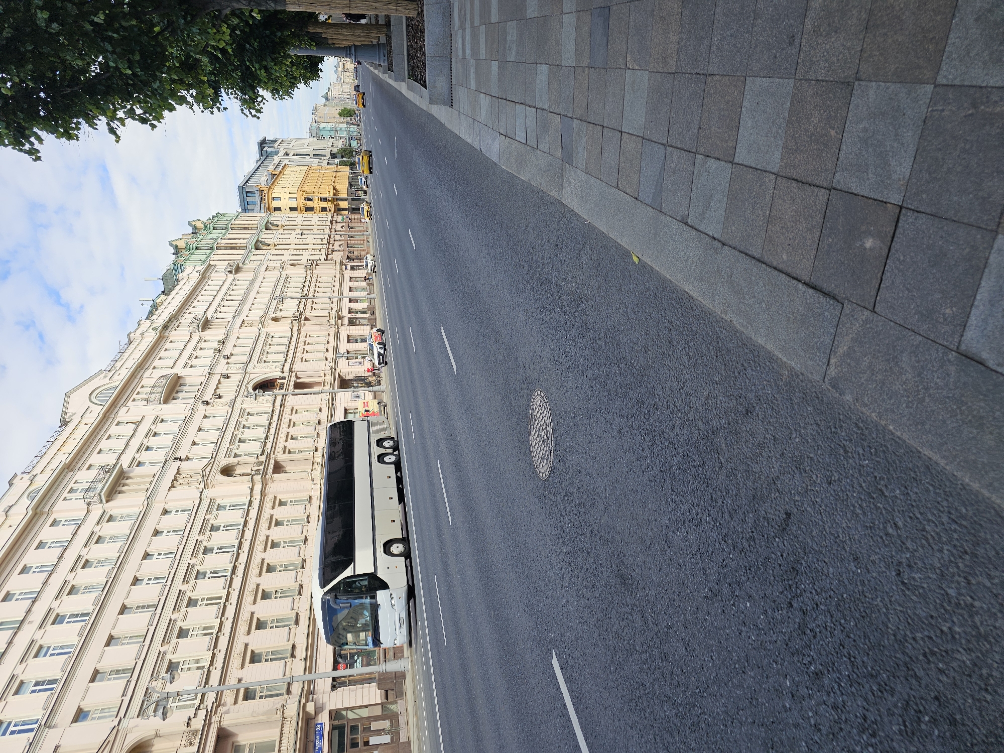 The image shows a street in Moscow, Russia, on a cloudy day. The street is wide and paved with asphalt, with white lane markings. On the left side of the street, there are several large, multi-story buildings with ornate architecture and many windows. A white tour bus is parked on the side of the road. On the right side of the street, there is a sidewalk made of stone pavers, lined with trees. Several yellow taxis are visible on the street, moving away from the camera. A few pedestrians are walking on the sidewalk. The overall scene depicts a typical urban streetscape in Moscow.
