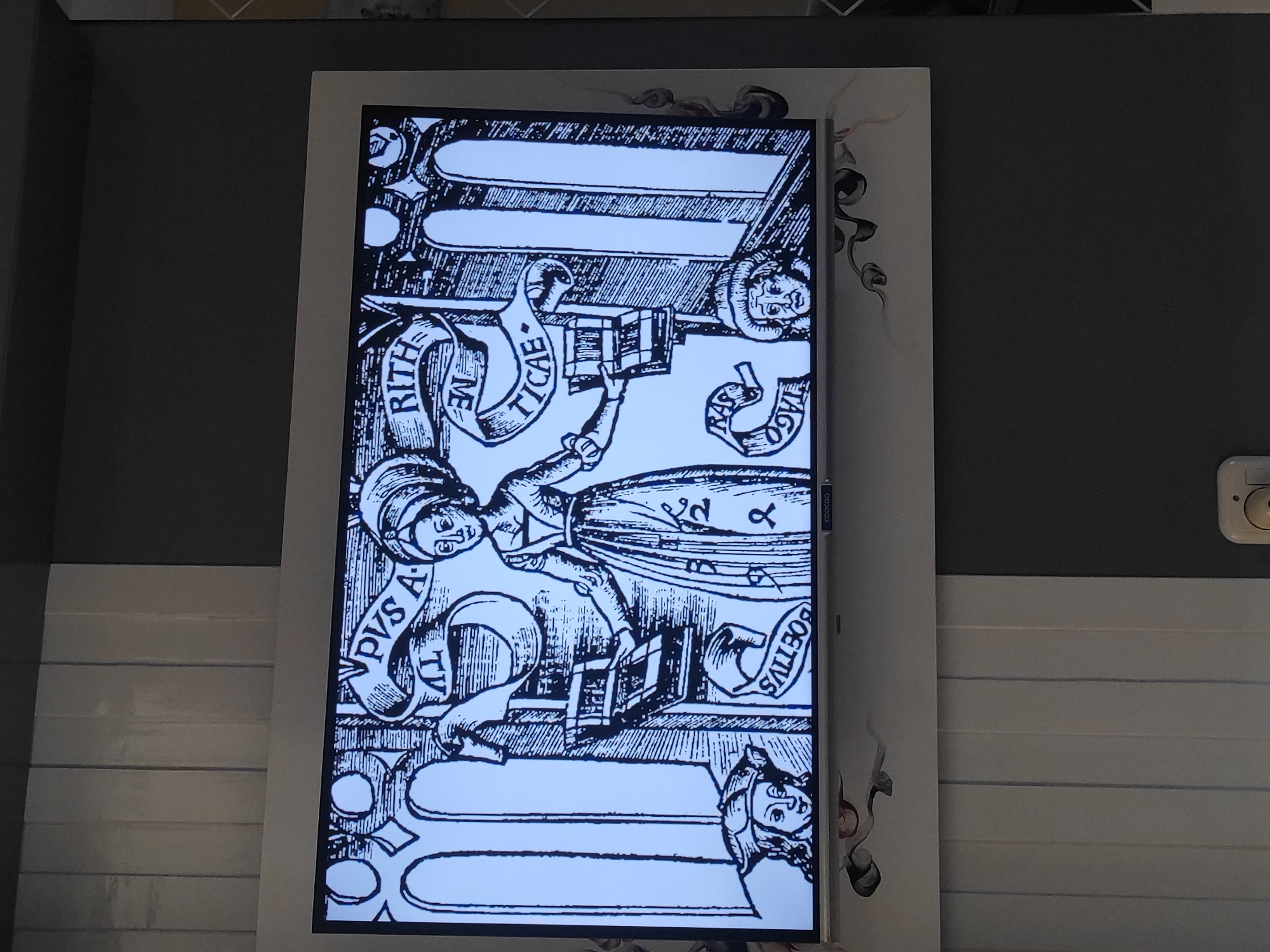 The image shows a television screen displaying a black and white illustration. The illustration features a central female figure in traditional attire, holding what appear to be books. She is surrounded by architectural elements and banners with text. Below her, on either side, are two smaller male figures, also in historical clothing. The television itself is framed by a white border and is mounted on a wall. The wall is painted in alternating vertical sections of light gray and dark gray. The lower part of the wall has a decorative design with swirling dark lines and floral motifs. This scene is located in the city of Sumedang, Indonesia.