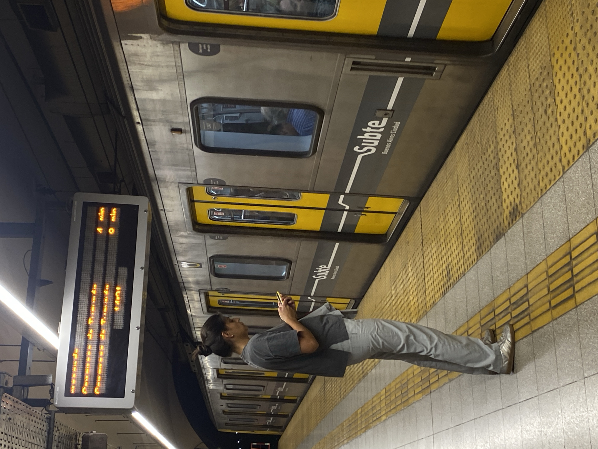 A woman stands at a subway platform in Buenos Aires, Argentina. She is looking at her cell phone. The train has the word "Subte" displayed on its exterior. An electronic sign displaying information is visible to the left of the woman. A yellow tactile paving edge is visible on the platform.