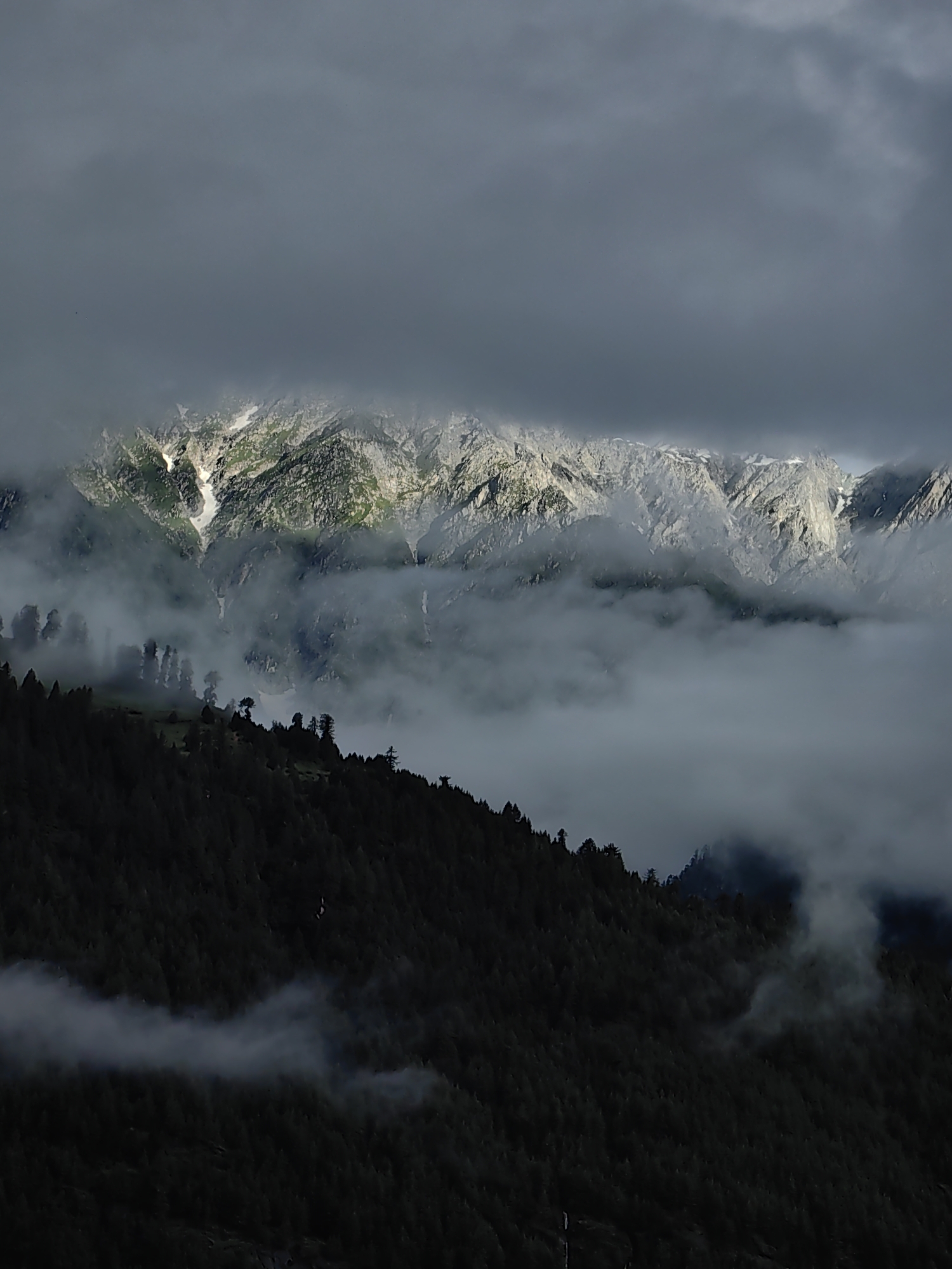 The image depicts a dramatic mountain landscape partially obscured by clouds and mist. The foreground is a dark, densely forested slope, likely pine trees, that recedes into the distance. A layer of thick, white mist hovers above this forest, creating a sense of depth and mystery. Behind the mist, the rugged peaks of the mountains are visible, their rocky surfaces catching the light. Patches of snow are present on the higher elevations, contrasting with the grey rock and green vegetation. The sky is filled with heavy, dark clouds, with shafts of light breaking through in places, illuminating sections of the mountain range. The overall impression is one of wild, untamed natural beauty. This scene is consistent with the mountainous terrain surrounding Vashisht, India, a village known for its hot springs and proximity to the Himalayas.