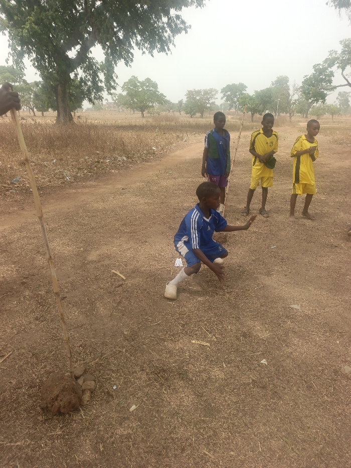 Four children stand in a dry, grassy field. One child in blue and white kneels, holding a white object, possibly a ball. Three other children stand behind him; one wears blue and the other two wear yellow soccer uniforms. In the foreground, a slender tree branch leans against a pile of earth. Trees dot the landscape in the background under a pale sky. A dirt path runs through the field.