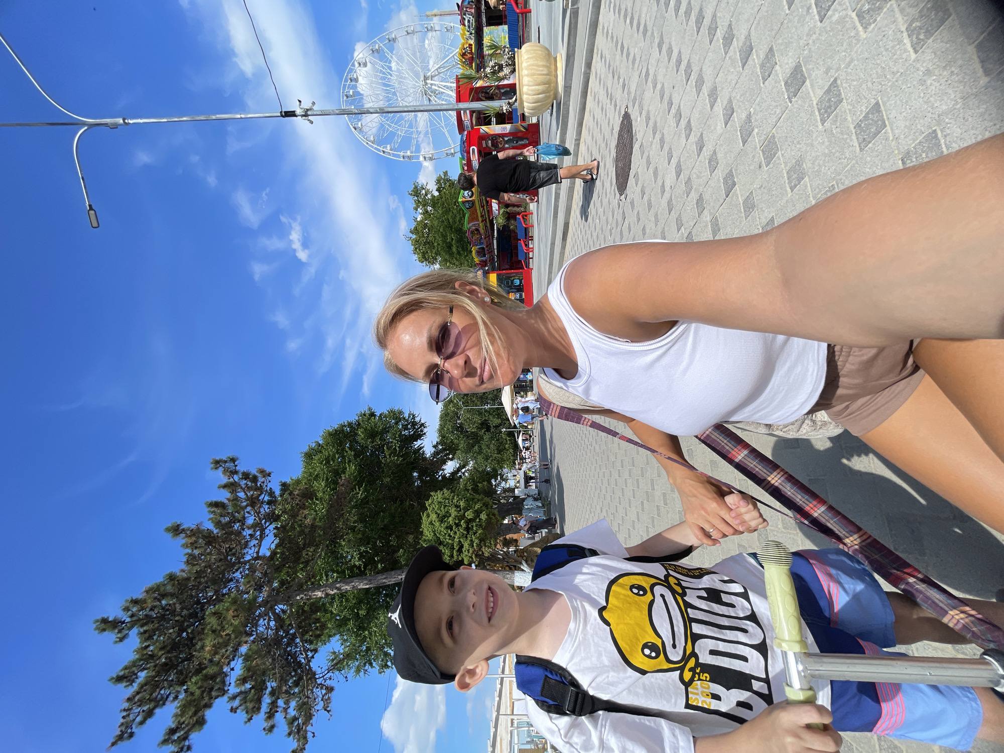 In Anapa, Russia, a woman and a boy stand on a paved promenade under a bright blue sky. The woman, with blonde hair and sunglasses, wears a white tank top and brown shorts. The boy, wearing a black cap, a white t-shirt with a cartoon duck, and blue shorts, holds a scooter. They are holding hands, and the woman is taking a selfie. In the background, a Ferris wheel and carnival rides are visible, along with trees and other people walking. The scene appears to be a sunny day in a seaside resort.