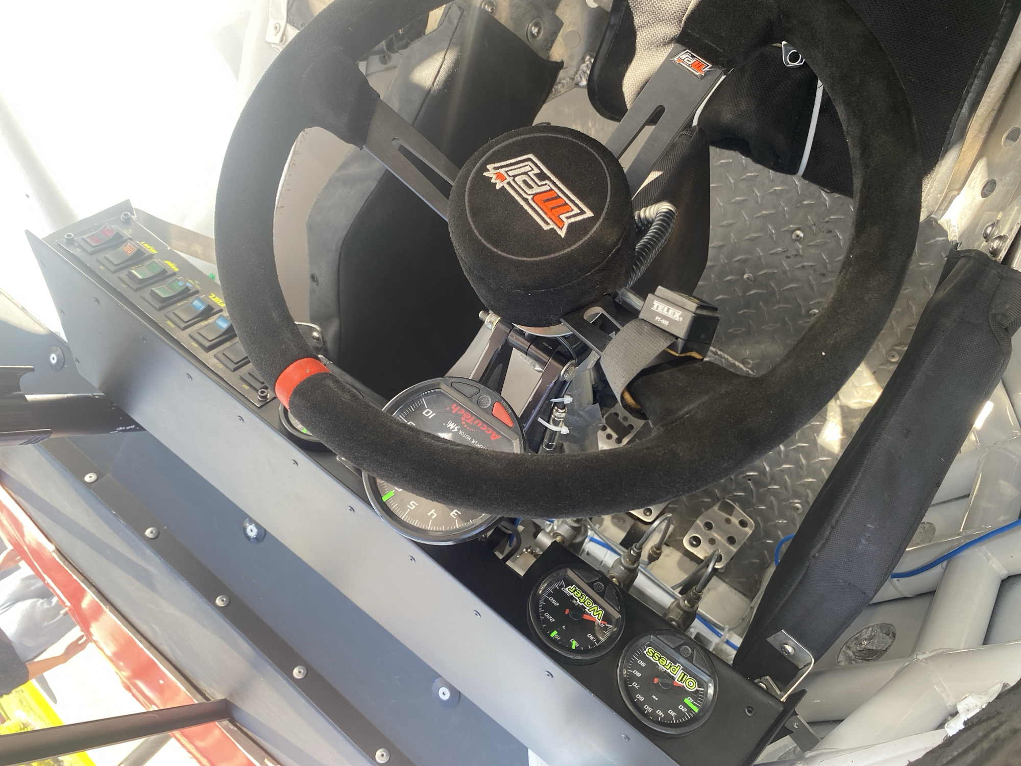 This is a close-up interior shot of a race car cockpit. The steering wheel, covered in a black, suede-like material with a red accent at the top, dominates the foreground. The steering wheel features a center hub with the MPI logo. Behind the steering wheel, the car's seat and safety harness are visible.
Various gauges and switches are mounted on a panel to the left of the steering wheel. The gauges display information such as oil pressure, water temperature, and engine RPM. The switches include labels like "FUEL". The pedals are also visible near the bottom of the frame.
The car's interior is made of metal, with a diamond-plate pattern on the floor. Blue and black wires are visible within the car. A Telex PT-300 device is mounted nearby the steering wheel.
The image does not offer any information to allow determining the location in the city of San Bernardino Tlaxcalancingo, Mexico.