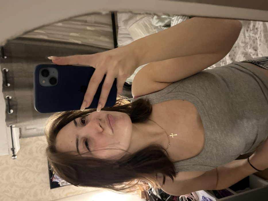A young woman takes a selfie in a mirror. She has brown hair, fair skin, and wears a grey tank top. A gold cross necklace hangs around her neck. She holds a dark blue iPhone in her left hand, her long nails visible. The background reveals a bedroom setting with curtains, bedding, and a glimpse of furniture. The image is slightly rotated.