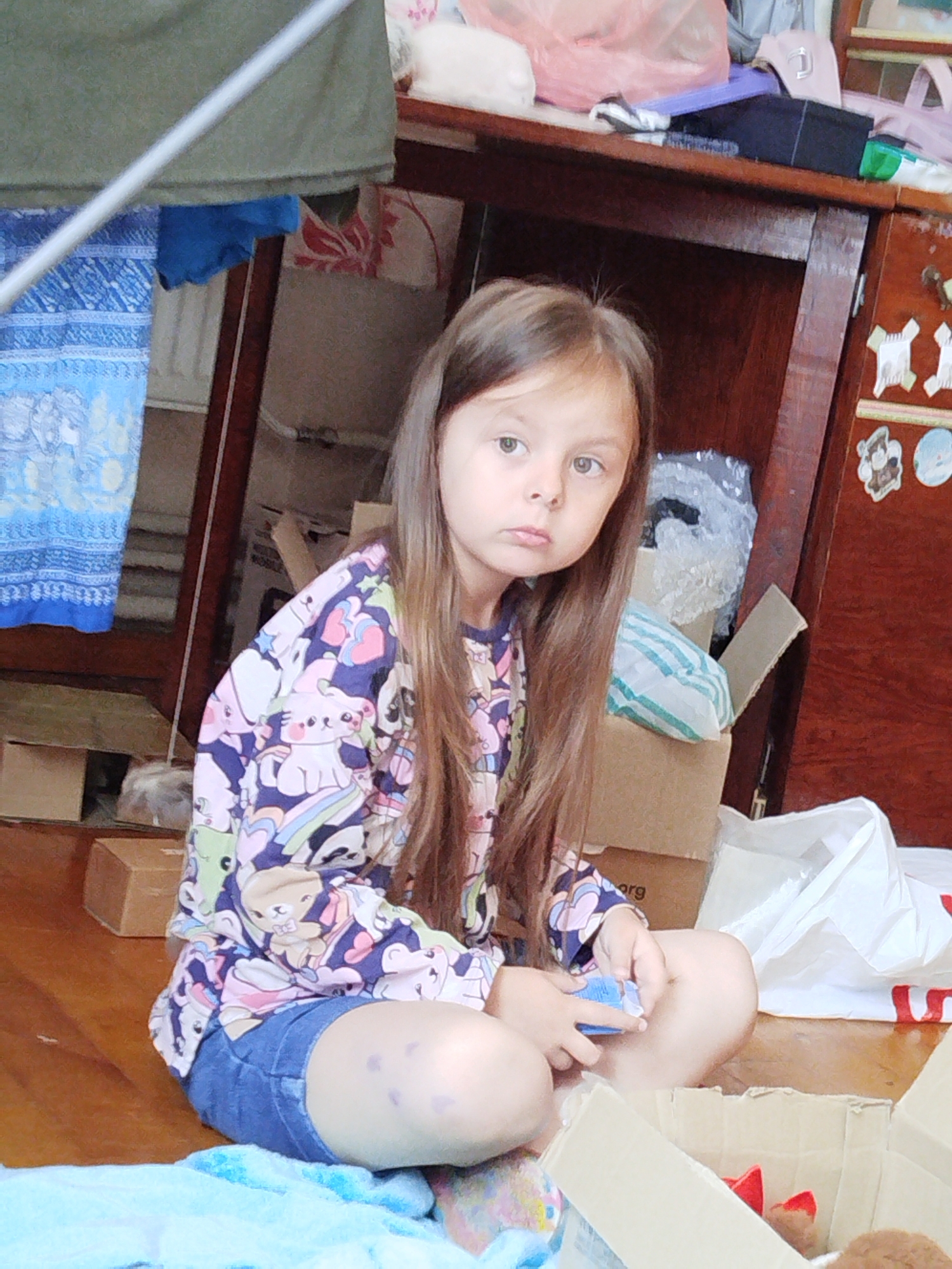 A young girl with long, brown hair sits cross-legged on a wooden floor. She is wearing a colorful long-sleeved shirt with cartoon animal prints and blue denim shorts. Her expression is pensive, and she is holding a small blue object in her hands. The room appears to be cluttered, with various boxes, bags, and what looks like laundry in the background. A wooden shelf unit with drawers is visible to the right, adorned with stickers. The lighting suggests it could be daytime, possibly indoors with artificial lighting. The overall impression is a candid shot of a child in a domestic setting, possibly in Vinnytsia, Ukraine.