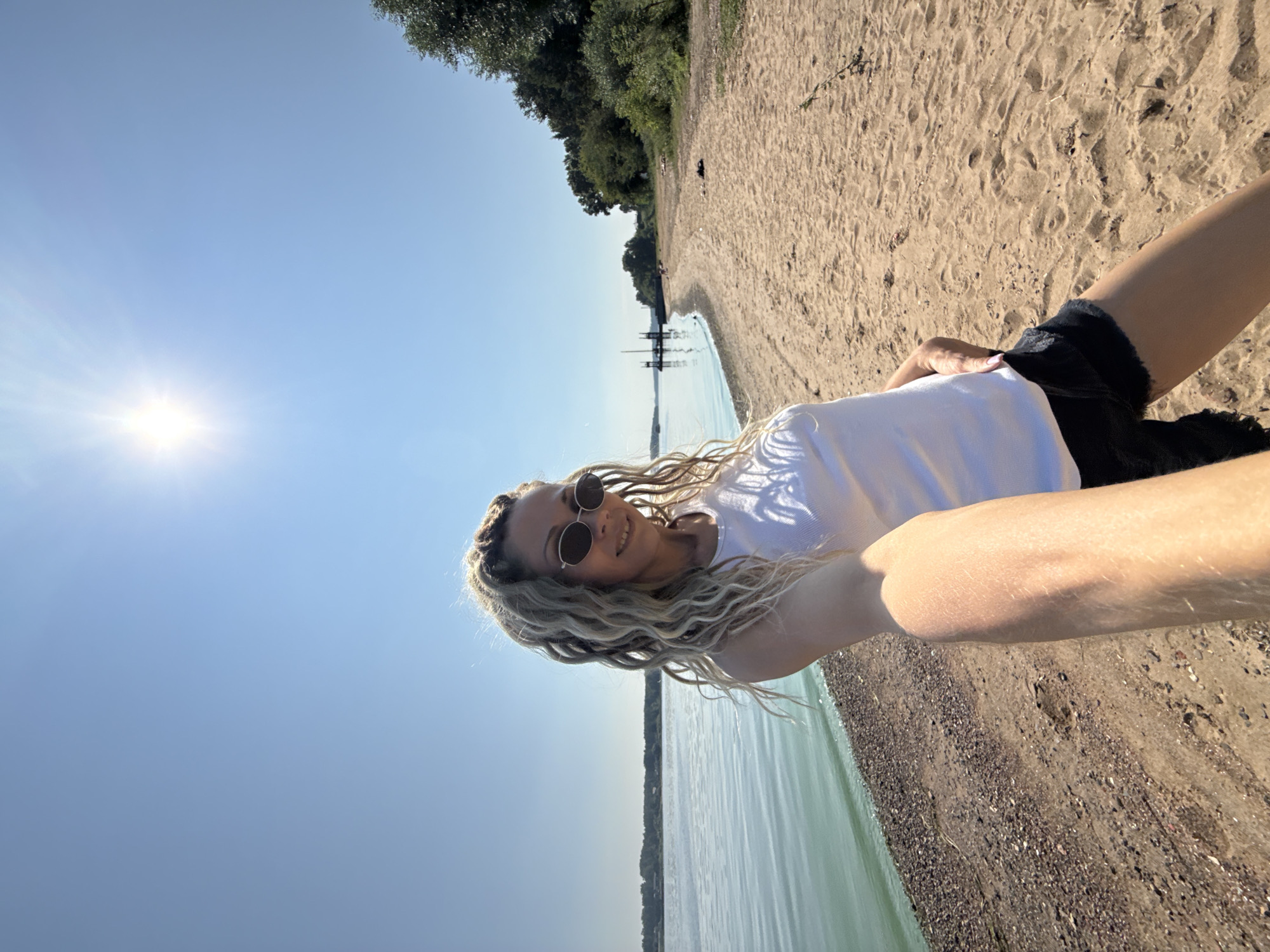 A woman with blonde dreadlocks and sunglasses is taking a selfie on a sandy beach next to a body of water. She is wearing a white tank top and black shorts. The sun is shining brightly in a clear blue sky. In the background, there is a dock with a boat, trees, and more beach. The scene appears to be in Rusanovo, Russia.