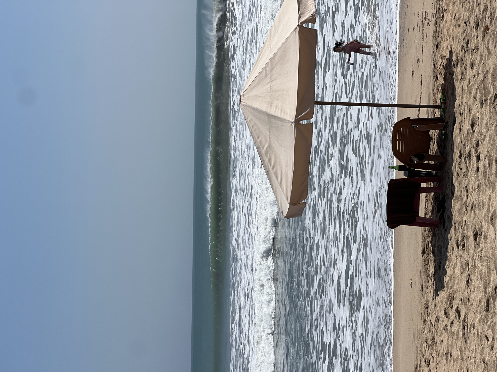 In Kuta, Indonesia, a beach scene unfolds under a clear, hazy sky. The sand, a light brown, is marked with footprints and a few shadows from unseen objects. In the foreground, a large, beige beach umbrella is open, casting a shadow on the sand. Beneath it, two plastic chairs, one red and one orange, sit near a small black table. A green bottle rests on the table.
In the middle ground, the ocean is a deep teal, with large, powerful waves breaking and churning white foam. The water's surface is a textured expanse of whitecaps and ripples, extending to the horizon. A young girl, wearing a pink swimsuit, stands in the shallow water at the edge of the sea, holding a bottle and looking towards the shore. She is partially obscured by the umbrella's pole and the breaking waves. The overall impression is one of a bright, sunny day at a popular tourist beach.
