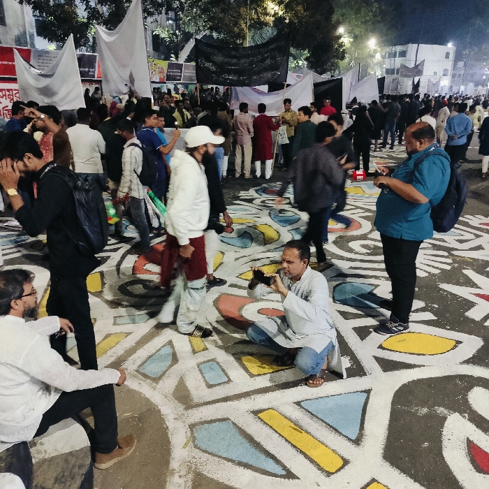 A large crowd stands on a street covered in geometric shapes painted in yellow, blue, red, and gray. People are taking pictures, some kneeling to get a better angle. Banners and flags are visible in the background, suggesting a gathering or protest. The light indicates it is evening or night. The crowd is diverse, with people wearing various clothing styles.