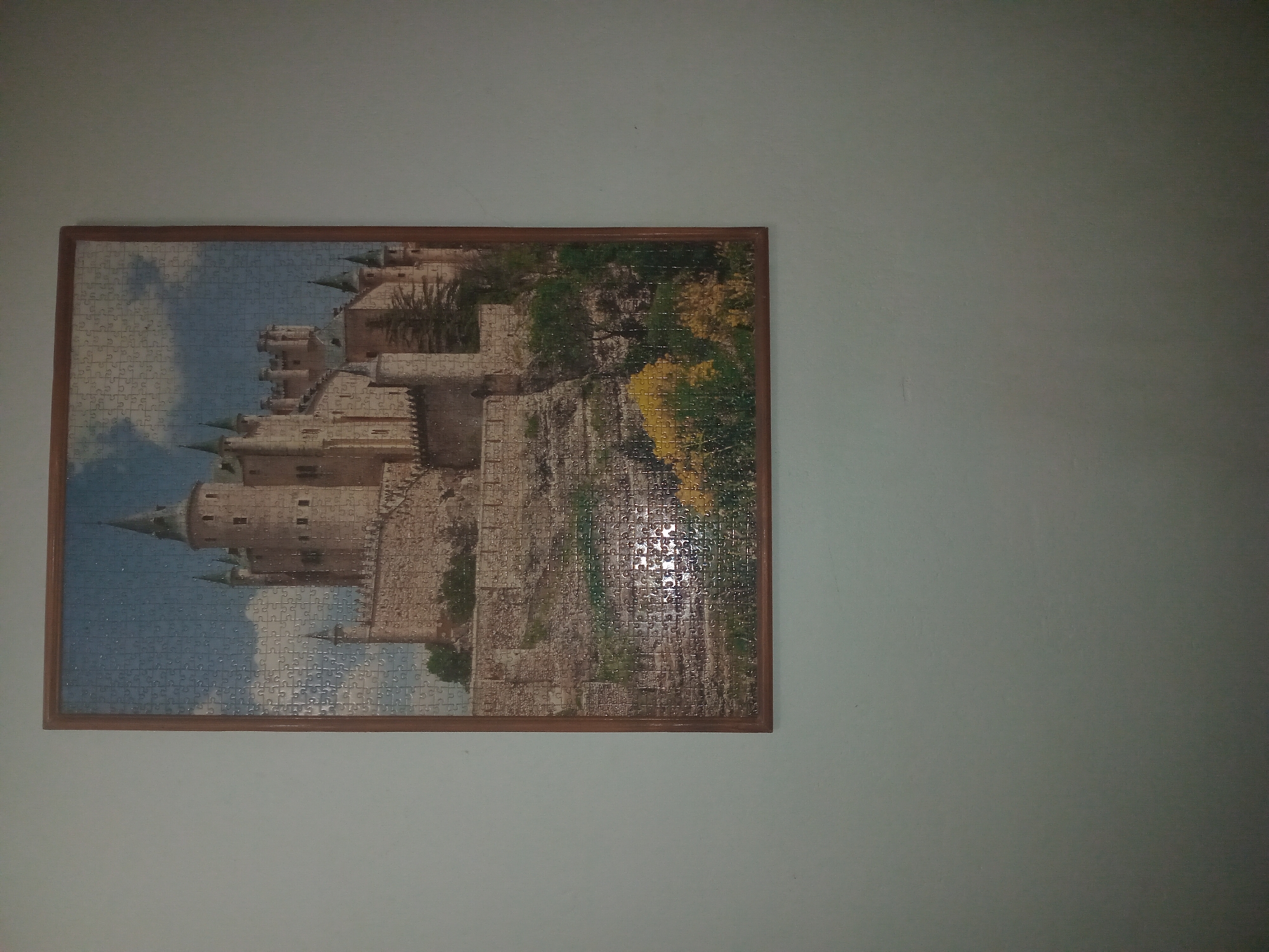 This is a medium shot, eye-level photograph of a framed jigsaw puzzle of a castle hanging on a light-colored wall. The puzzle depicts a large stone castle with multiple towers and spires, set against a blue sky with white clouds. The castle appears to be on a hill, with stone walls and terraces leading up to it. There are trees and some yellow flowers visible in the foreground. The frame of the puzzle is made of dark wood. The wall surrounding the puzzle is a plain, light, neutral color. The image does not provide any information about the city of Perechyn, Ukraine, as it is a photograph of a puzzle, not a depiction of a real-world location.