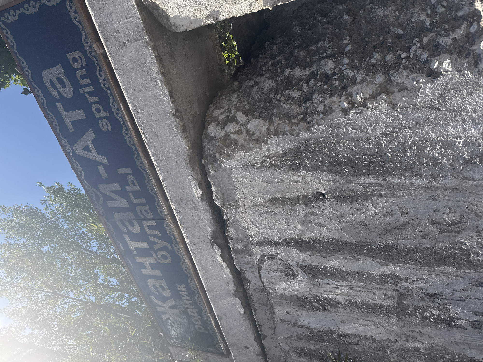 A sign on a stone structure reads "Жантай-Ата spring". The sign is dark blue with light-colored text and decorative borders. The text appears to be in a Cyrillic script, with "spring" written in English below the Cyrillic. The sign is mounted on a rough, grey concrete structure that is cracked and weathered. Behind the sign, green trees and a bright blue sky are visible, with sunlight filtering through the leaves. The image appears to be taken from a low angle, looking up at the sign and the concrete structure. The context of Cholok, Kyrgyzstan is not directly evident from the image itself, but the text on the sign suggests a location of natural significance.