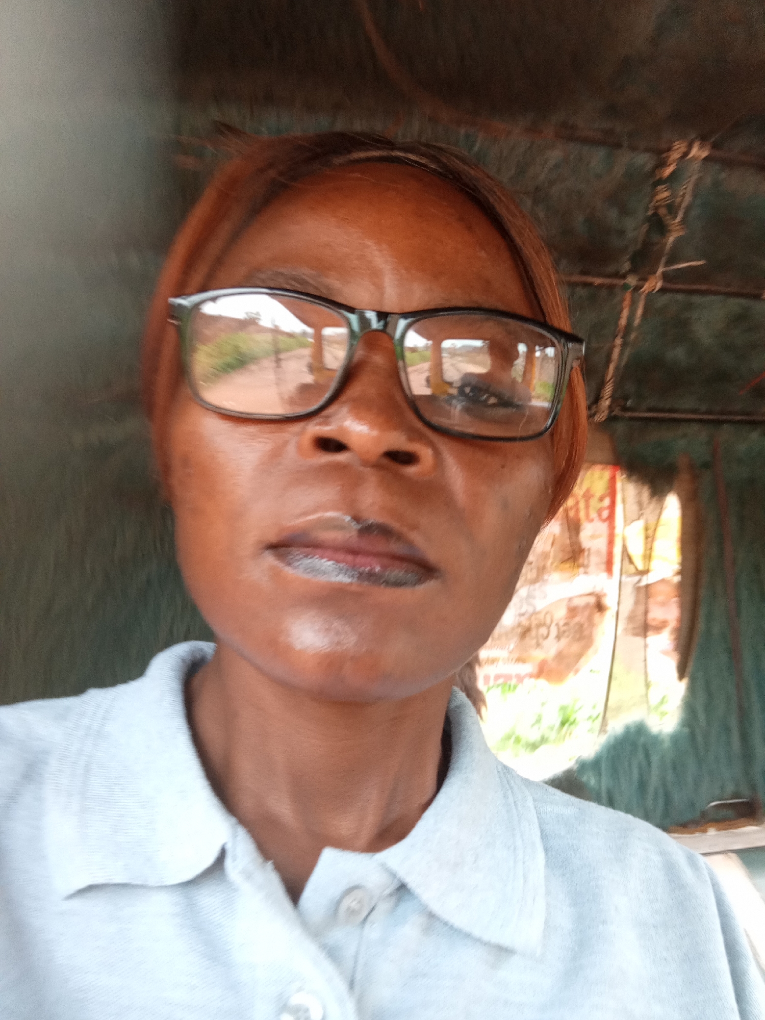 The image is a close-up, front-facing selfie of a woman wearing glasses and a light blue collared shirt. She has dark skin and reddish-brown hair. The background appears to be a rustic, enclosed space with some textured material on the walls and ceiling. There are glimpses of the outdoors through an opening, showing foliage and a dirt road. A reflection in her glasses shows a vehicle, possibly a tuk-tuk. The overall lighting suggests it is daytime, and the image appears to be taken outdoors or in an open-air structure. It is not possible to determine the specific location within Jalingo, Nigeria from the provided image.