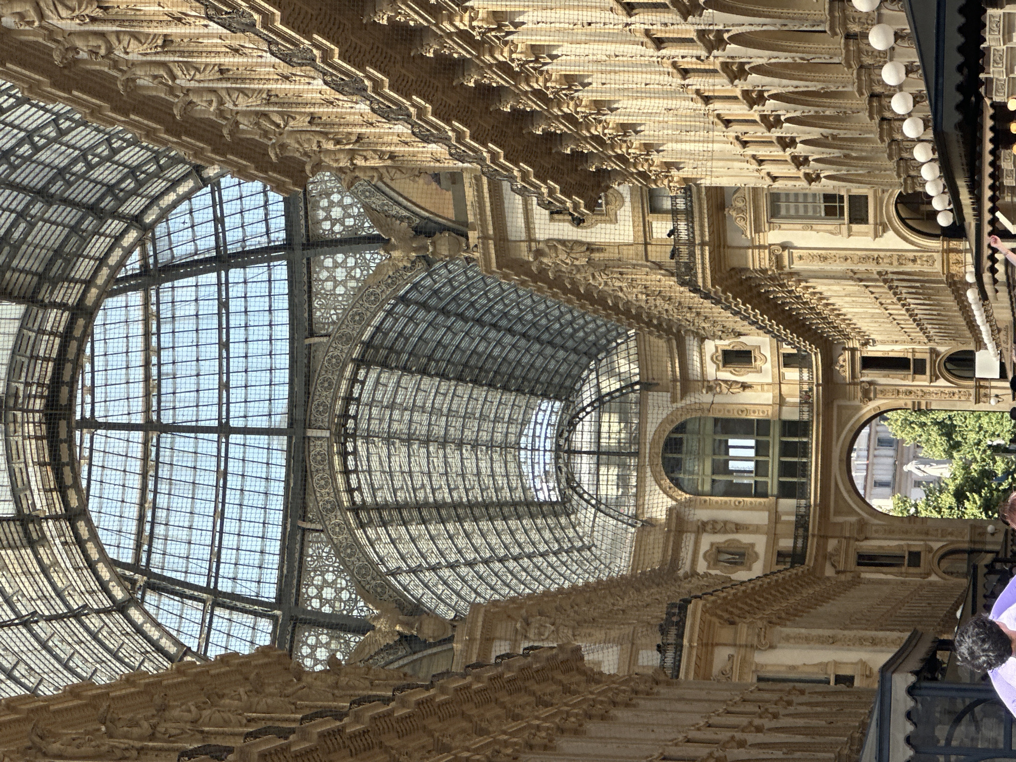 The image captures the Galleria Vittorio Emanuele II in Milan, Italy. This historic shopping arcade features an ornate glass and iron roof, supported by intricate metalwork and arches. The walls of the arcade are lined with elaborate stonework, including columns, statues, and balconies. Sunlight streams through the glass ceiling, illuminating the interior. In the foreground, the tops of awnings and some people are visible, suggesting a public space with commercial activity. Through an archway further down the arcade, a statue and trees can be seen, indicating an outdoor area beyond. The overall impression is one of grandeur and architectural splendor.