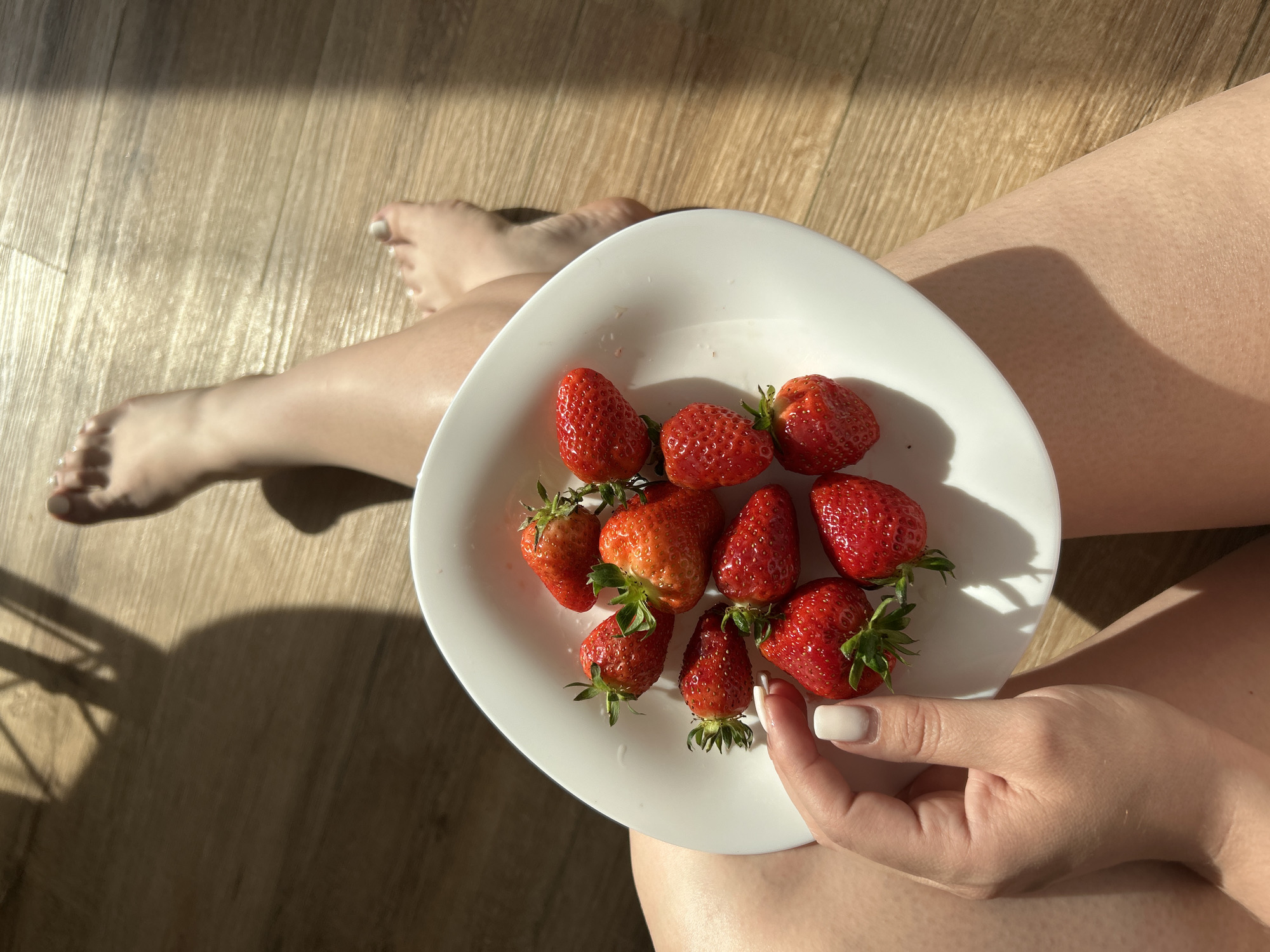A top-down shot shows a woman sitting on a light brown wood floor, legs crossed, holding a white plate filled with approximately ten strawberries. The woman has light skin and manicured fingernails with a white polish. One foot is visible near the top left of the frame. Light and shadow suggest a sunny day. There is no indication of the city of Chaikovsky, Russia.