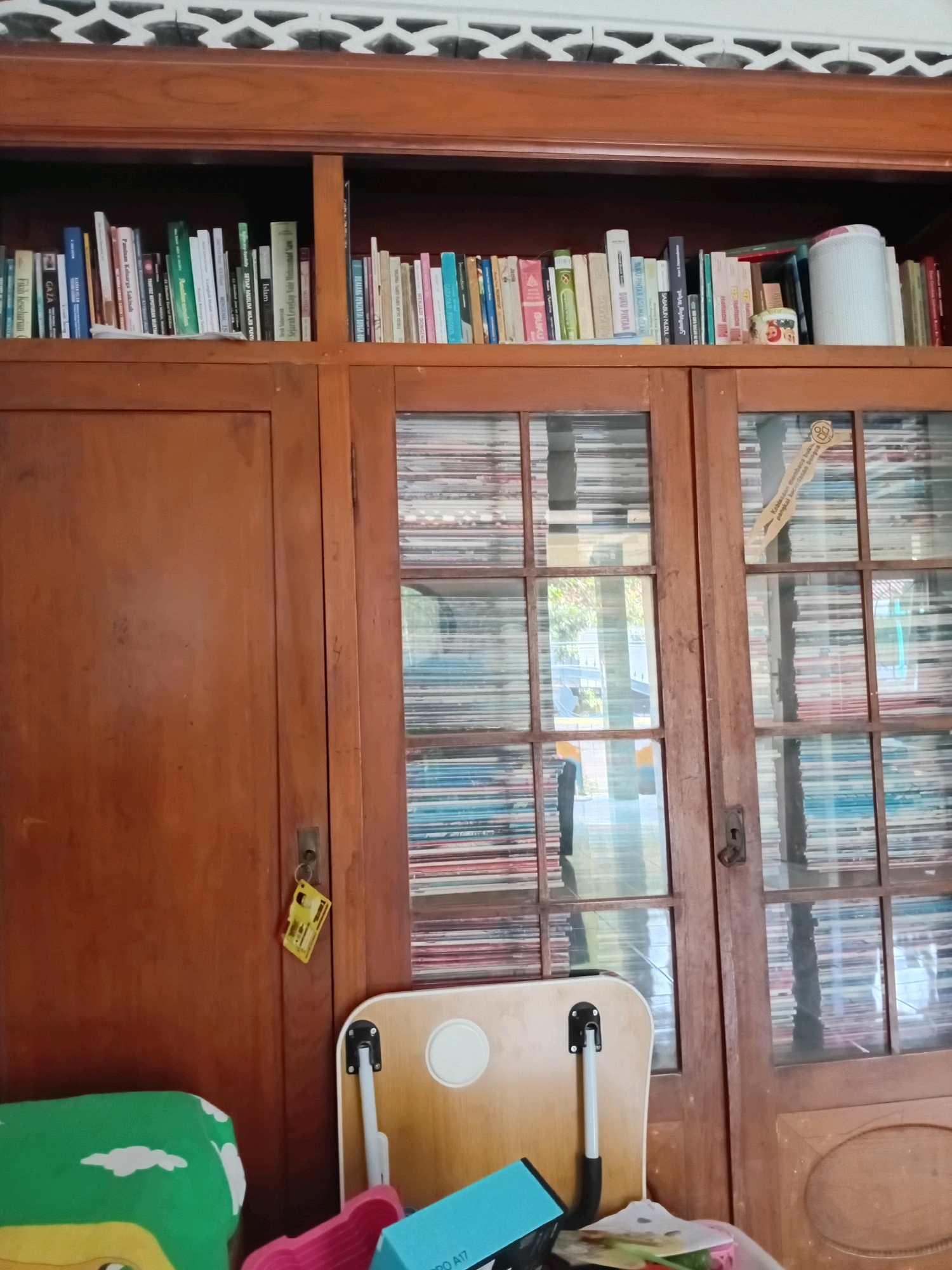 The image shows a wooden bookshelf with several books. The bookshelf is divided into two sections. The left section is a closed wooden door, while the right section has glass doors revealing more books. The top of the bookshelf has a decorative wooden trim. In front of the bookshelf, there is a small, foldable wooden table with white legs, a green pillow with a white cloud pattern, and a pink object that appears to be a holder or basket. There is also a blue object, possibly a phone, and some papers on the floor. The overall scene suggests an indoor environment, likely a room in a residential or public space. The mention of Gebang, Indonesia implies the geographical context of the image.