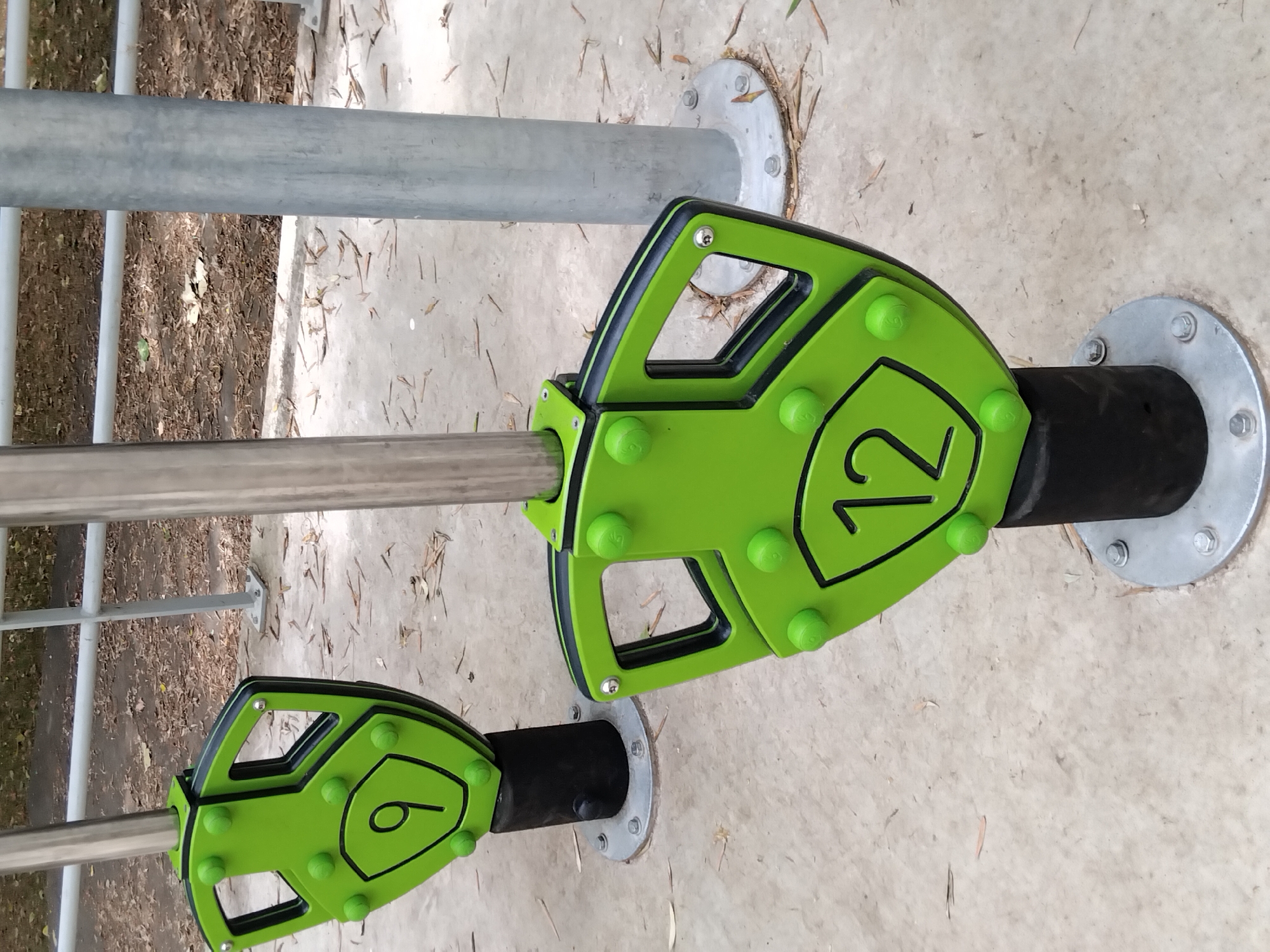 The image shows two green exercise equipment components, each attached to a vertical metal pole. The component in the foreground is a shield-shaped piece with the number "12" prominently displayed in black. It has several green spherical knobs on its surface and black trim around its edges. This component is mounted on a black cylindrical base. In the background, a similar shield-shaped component with the number "9" is visible, also attached to a metal pole and mounted on a black base. Both bases are secured to the ground with metal plates and bolts. The ground surface is concrete, with scattered dry leaves and debris. Behind the equipment, a metal railing is visible, and beyond that, a grassy area with trees. The scene appears to be an outdoor exercise park. The image does not provide enough information to determine the specific location in Vila Nova de Famalicão, Portugal.