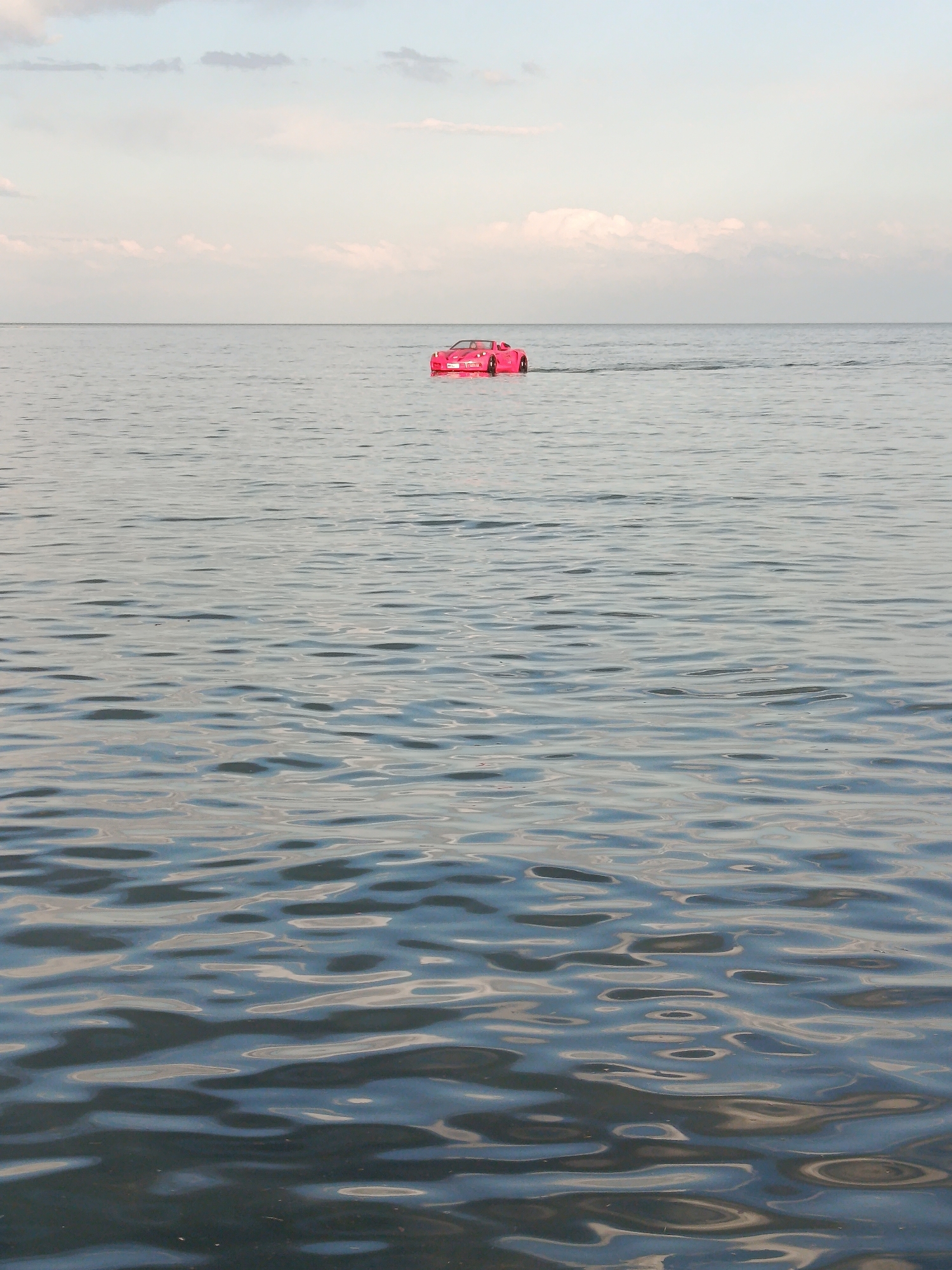 A pink sports car is driving on the surface of a large body of water. The car is positioned in the upper middle of the frame. The water is calm with gentle ripples, reflecting the soft colors of the sky. The sky is a pale blue with scattered white clouds, suggesting it is either early morning or late afternoon. The horizon line is visible in the distance, separating the water from the sky. The overall scene is surreal and unusual, with the car appearing to float or drive on the water. There are no people visible in the image.