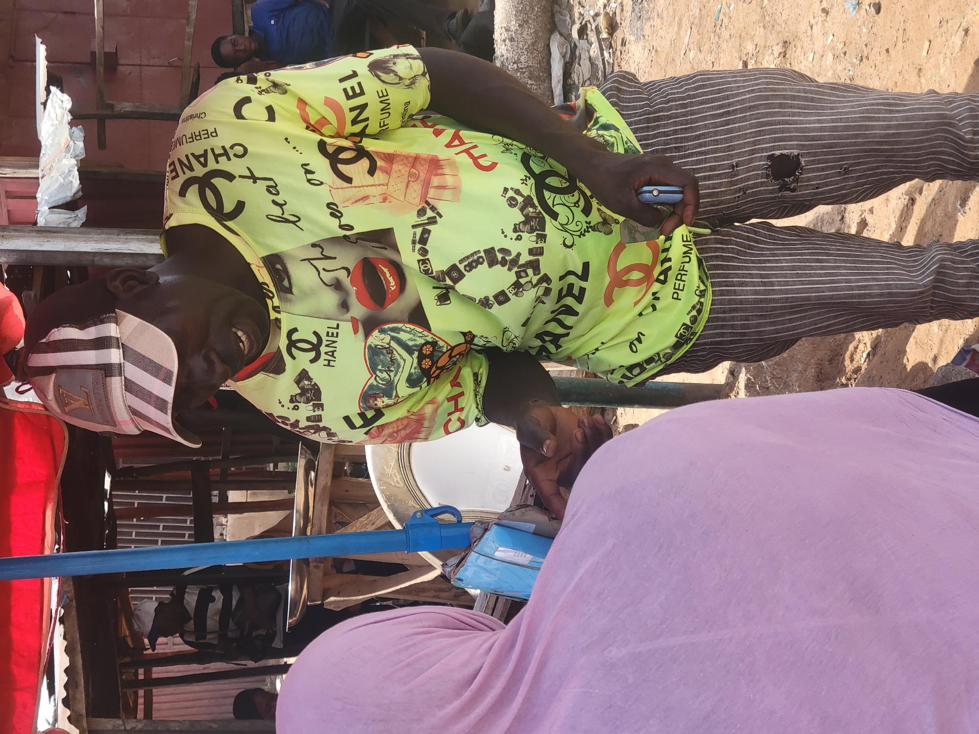 The scene shows a man in Kwamba, Nigeria, interacting with a person wearing a pink hijab. The man wears a yellow Chanel-patterned shirt, horizontally striped pants with a hole, and a patterned cap. He holds a mobile phone in one hand. In the background, there is a partially constructed wooden structure and some visible urban elements. The ground appears to be sandy or dusty.