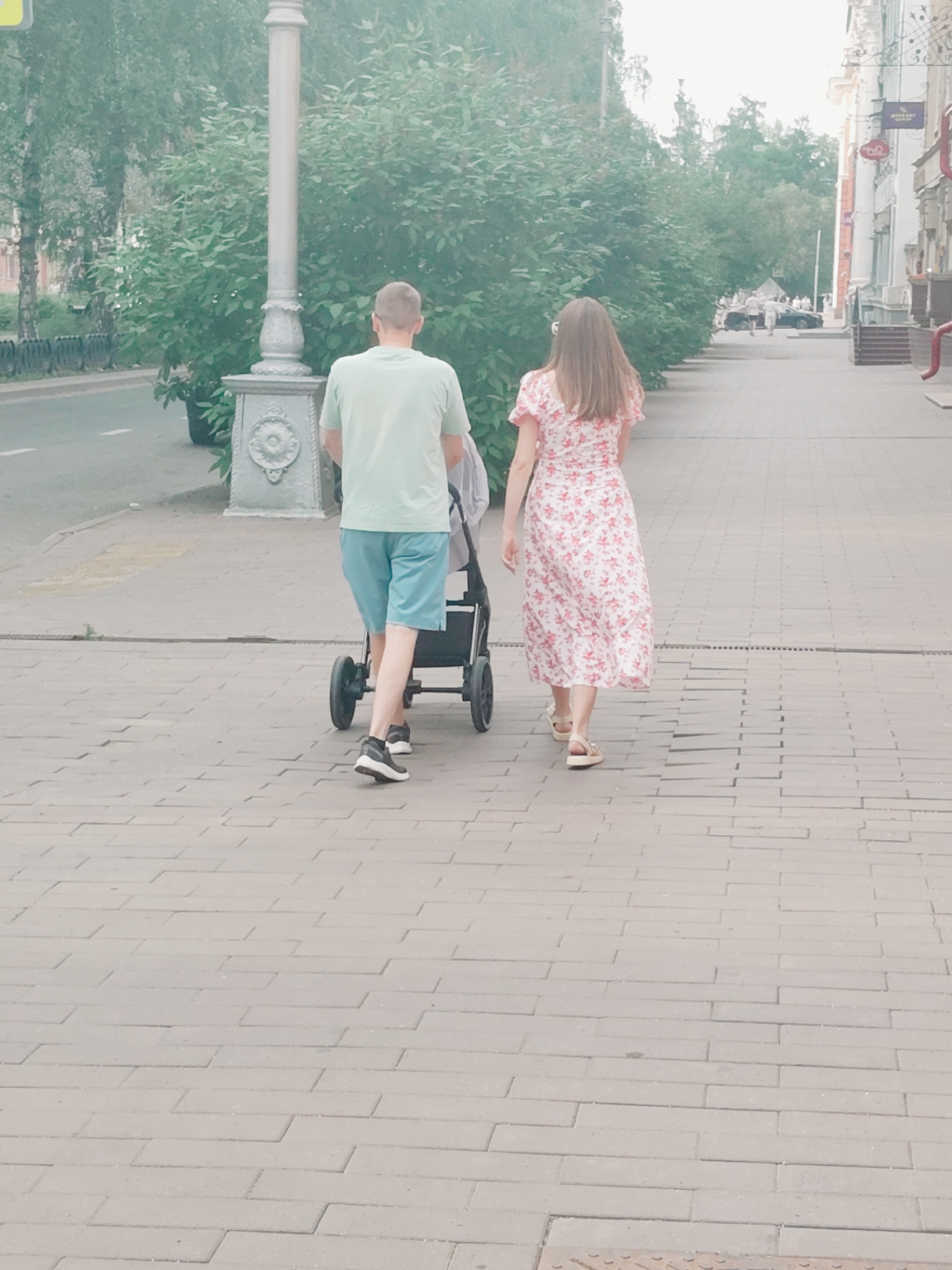 Two individuals, a man and a woman, are walking along a pedestrian sidewalk paved with light-colored rectangular bricks. The man, wearing a light green t-shirt and blue shorts, is pushing a stroller. The woman, in a floral print dress, walks beside him. They are walking away from the camera, down a street lined with green trees and buildings. The scene suggests a casual stroll in a city environment, possibly in Kemerovo, Russia, given the architectural style of the buildings and the overall atmosphere. The lighting is soft, indicating it might be daytime, possibly overcast or early morning/late afternoon.