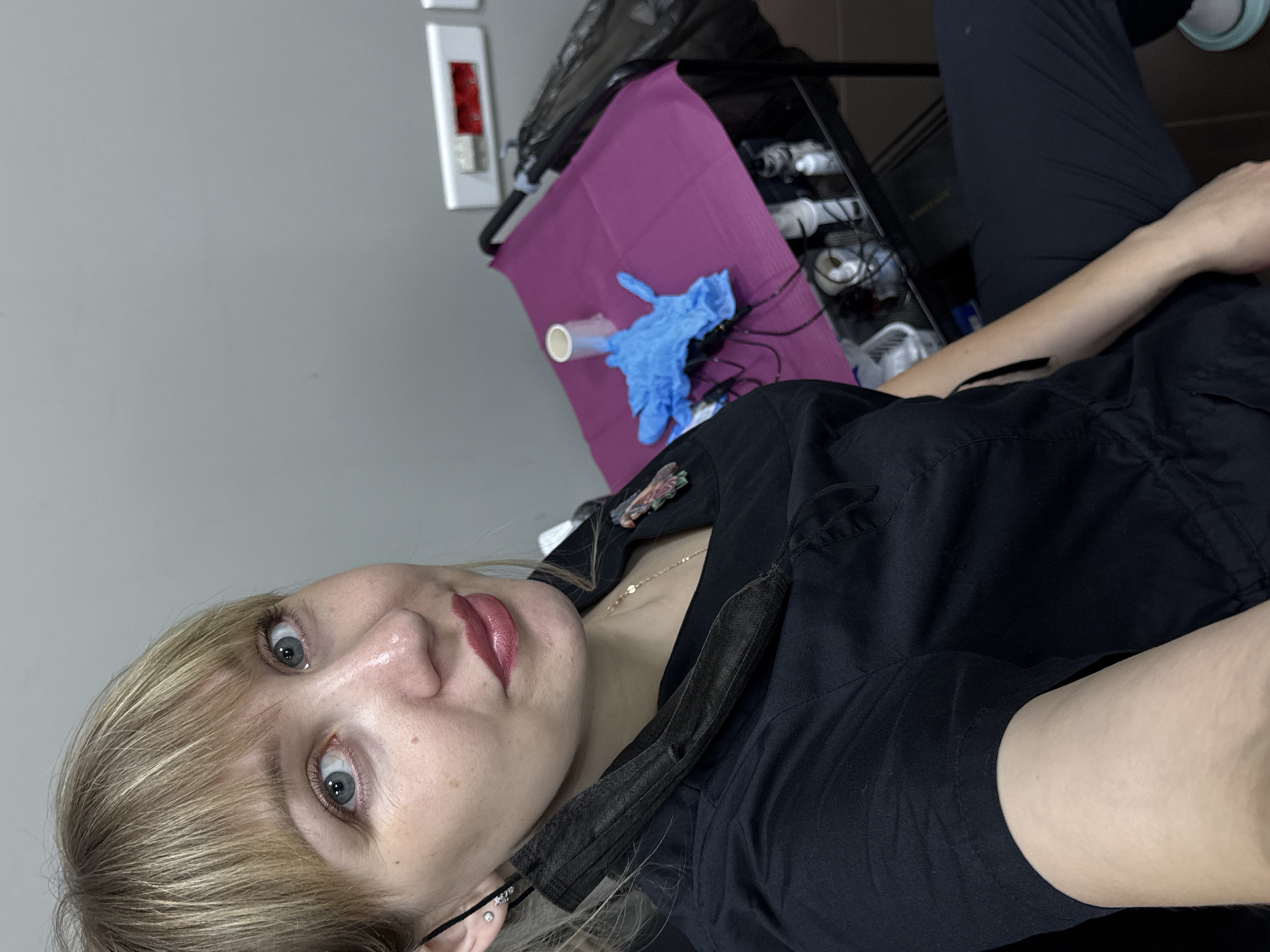 A woman with blonde hair and blue eyes, wearing black scrubs and a black face mask hanging from her ear, is taking a selfie. She has pink lipstick on and a silver earring in her left ear. In the background, a purple cloth covers a rolling cart with various supplies, including blue gloves, a roll of paper, and bottles. The wall behind her is light gray, and there is an electrical outlet on it. The floor is tiled. The location is identified as Chaikovsky, Russia.