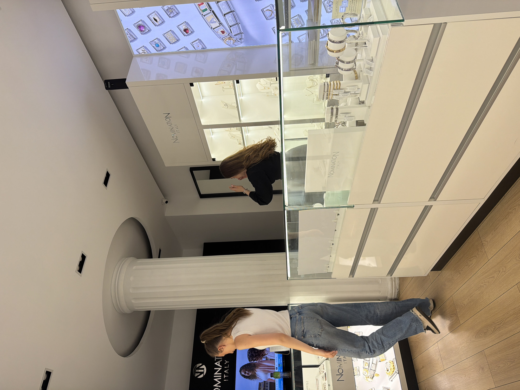 The image captures the interior of a brightly lit Nomination Italy jewelry store, likely located in Sofia, Bulgaria. The perspective is from an elevated angle, looking across the retail space.
Two women are visible. On the right side of the frame, a woman with long brown hair, dressed in a black top, is leaning into a glass display case, closely examining its contents. Her reflection is cast in a mirror situated behind the display. Towards the left, another woman with brown hair, tied back, wearing a white t-shirt and blue jeans, stands facing a different display case, looking down at the showcased items.
The store is furnished with modern, white display cabinets featuring glass tops and fronts. These cases are filled with various jewelry pieces, including bracelets, rings, and charms, illuminated by internal lighting. The "NOMINATION ITALY" logo is clearly visible on multiple display cases and possibly on a digital screen in the background. The flooring consists of light-toned wooden planks.
A prominent architectural feature is a large, fluted white classical column with a circular base and capital, extending from floor to ceiling in the central left area of the image. The ceiling is white and incorporates recessed lighting fixtures. A small black security camera is mounted high on a wall. The repeated brand name "NOMINATION ITALY" is evident, and faint Cyrillic text can be observed on some product labels or informational signs within the display cases, supporting the location context of Sofia, Bulgaria. The scene depicts a typical retail environment during what appears to be business hours.