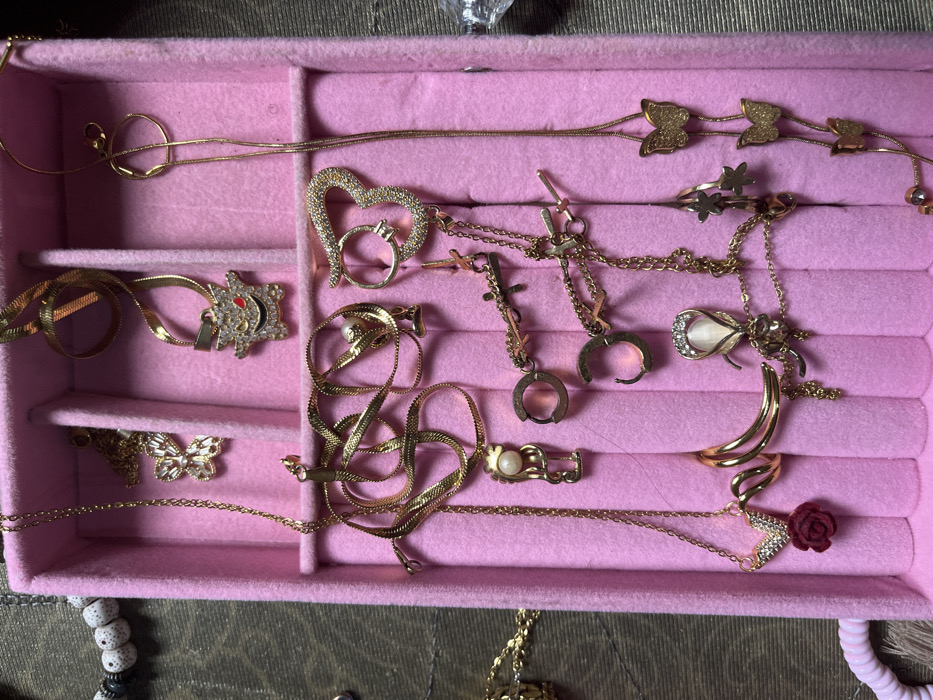 The image displays a pink, felt-lined jewelry organizer containing various gold-colored pieces, situated in the city of Ipetumodu, Nigeria. The organizer features multiple compartments.
**Objects within the organizer:**
- **Necklaces:** Multiple gold-colored necklaces are present. One necklace features a butterfly pendant encrusted with clear stones. Another has a Gengar-shaped pendant, detailed with clear stones and red eyes. A different necklace displays two brushed gold-colored butterfly pendants. Further necklaces include a pendant of a stylized figure holding a white pearl, a large heart-shaped pendant covered in clear stones, a red rose pendant beneath a V-shaped element with clear stones, an oval "cat's eye" stone pendant framed by clear stones, and a long chain with multiple small brushed gold-colored butterfly pendants and small spherical beads. Two necklaces feature small cross-shaped pendants.
- **Earrings:** Two gold-colored hoop earrings are visible in one compartment.
**Surrounding Environment:**
- To the immediate left of the organizer, a segment of a beaded bracelet with white, textured spherical beads and dark spacers rests on a green fabric surface.
- To the right of the organizer, a brown fabric surface is visible, upon which a small, clear, irregularly shaped object is positioned.