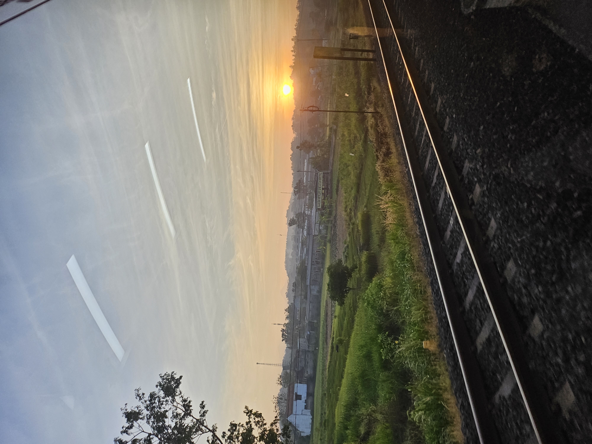 The image depicts a train track running diagonally through a rural landscape at sunrise, likely in the city of Margosari, Indonesia. In the foreground, the dark train tracks with ballast are visible. Beyond the tracks, a verdant green field stretches out, interspersed with a lone tree. Further in the distance, a cluster of buildings, possibly houses and a factory or industrial complex, are nestled amongst more trees and rolling hills. The sky is a soft gradient of pale blue and orange, with the sun a bright orb just above the horizon, casting a warm glow over the scene. Several horizontal white streaks, possibly reflections on a window or light trails, are visible in the sky. A dark tree branches into the frame from the upper left corner. The overall atmosphere is peaceful and serene, capturing the quiet beauty of a new day in the countryside.