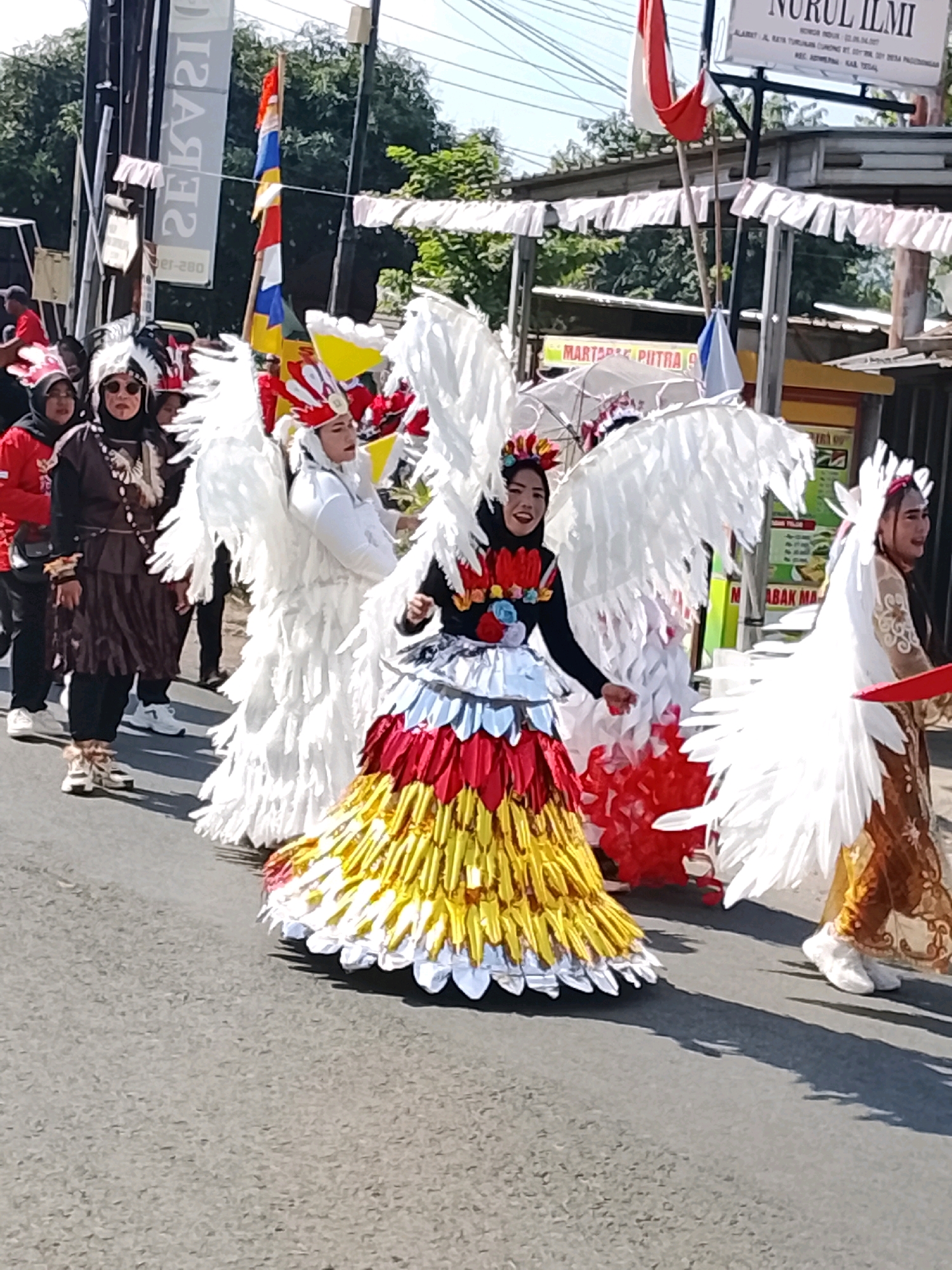 In Ujungrusi, Indonesia, a procession features individuals in elaborate costumes. The primary focus is on a group of participants adorned with large, white feather wings, reminiscent of angels or mythical birds. One individual in the foreground wears a black top with a colorful, layered skirt in red, yellow, and silver, also featuring wing-like embellishments. Another person to their left wears a white costume with white feather wings and a red and yellow headdress. Further left, a person in a dark brown outfit with a headdress and feather adornments walks alongside. The procession is taking place on an asphalt road, with buildings and trees visible in the background. Decorative white streamers hang across the road above the procession. Flags, including the Indonesian flag, are also visible. The overall scene suggests a cultural event or parade.