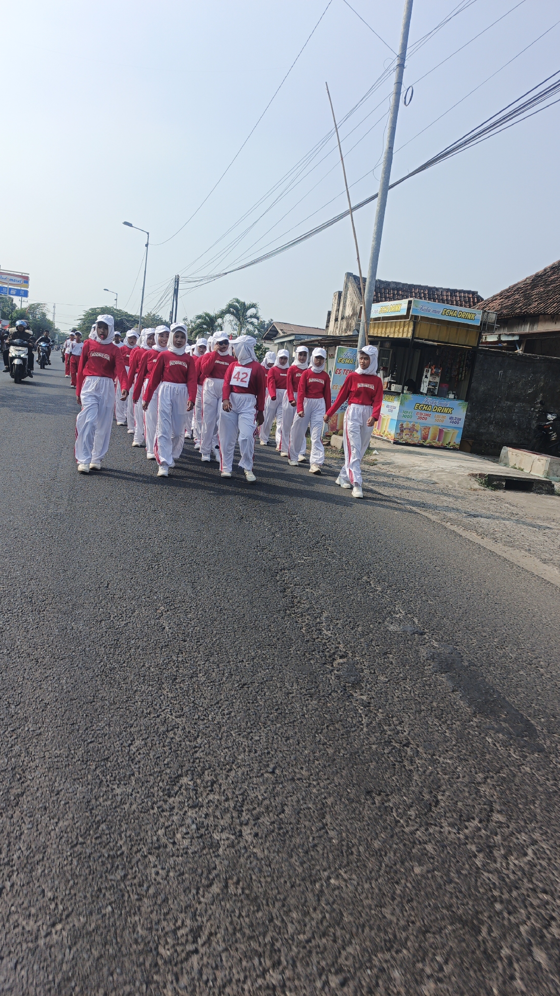 A group of people, likely students, are marching in single file on a paved road in Tampungrejo, Indonesia. They are all wearing white caps and red long-sleeved shirts with "INDONESIA" printed on them in white. Most are also wearing white pants. One person in the middle of the group has the number "42" displayed on their pants. They appear to be participating in a parade or some organized event. The road is asphalt and shows some wear and tear. To the right of the road, there are buildings and a cart selling drinks. In the background, there are utility poles with many overhead wires. The sky is clear and bright.