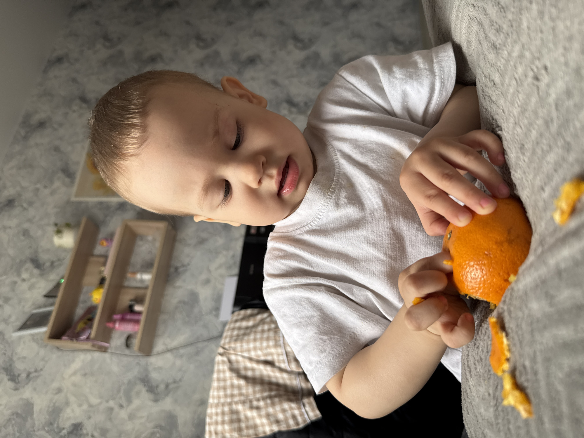 A young child with light blonde hair, wearing a white t-shirt, is peeling an orange. The child is seated on a grey sofa, with pieces of orange peel scattered nearby. In the background, there's a shelf with various items on it, including what appears to be art supplies, and a patterned blanket. The photo was taken indoors. There is no information indicating that this photo was taken in Rusanovo, Russia.