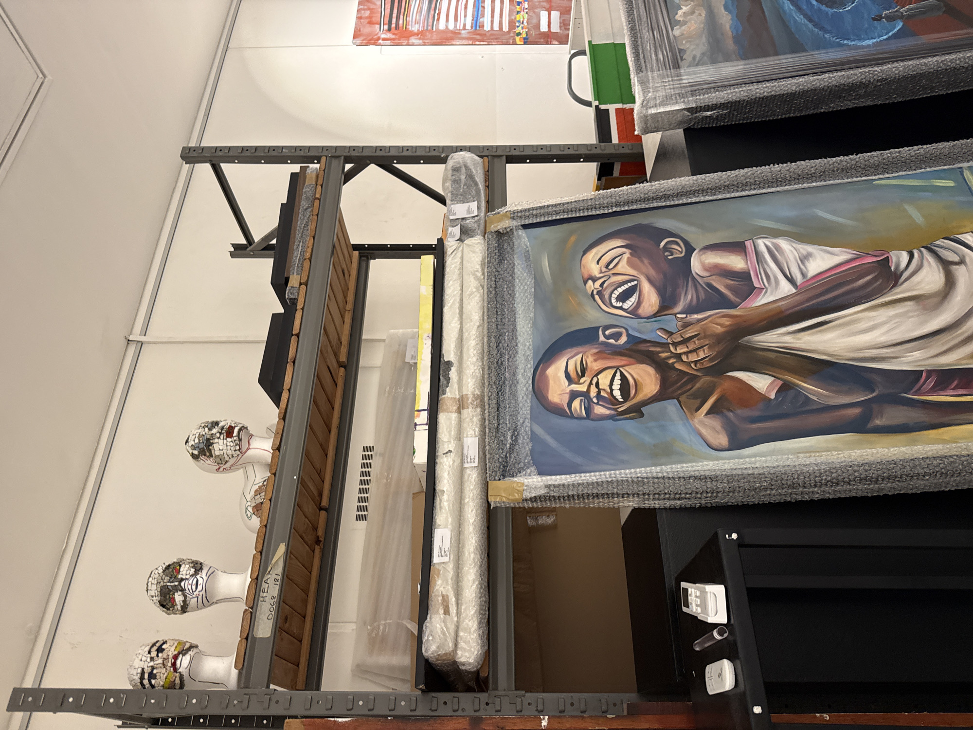 The image shows an interior storage space, possibly in an art storage facility, with artwork arranged on metal shelving. The artwork includes framed paintings wrapped in bubble wrap, wooden panels, and decorative mannequin heads. One painting prominently features two figures laughing. Other paintings are visible in the background. The exact location within Cape Town, South Africa, cannot be determined from the image.