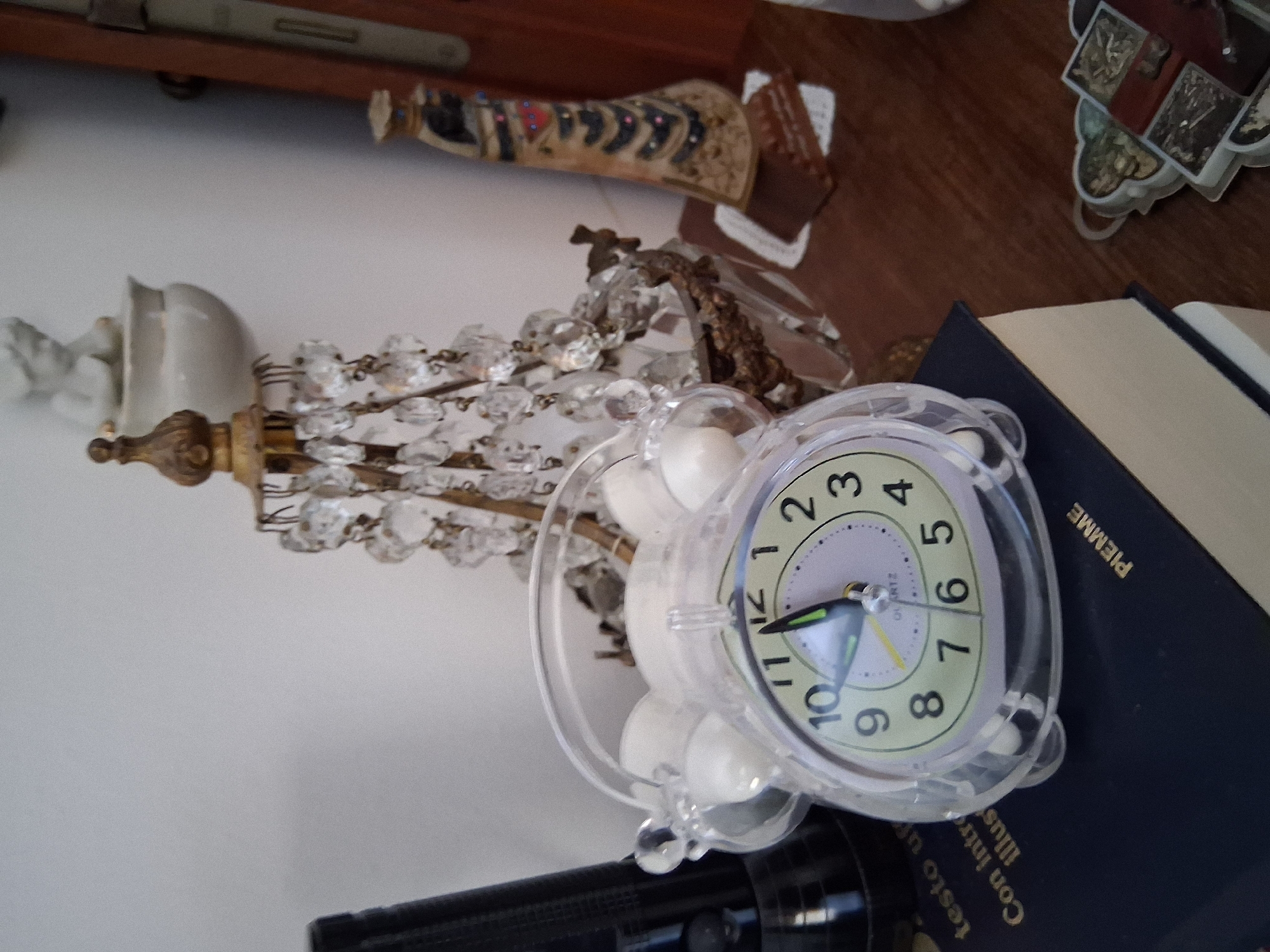 A close-up, eye-level shot captures a still life arrangement on a wooden surface, likely a table or shelf, within an interior. The foreground is dominated by a transparent, round alarm clock with white accents, its face displaying the time as approximately 10:56. The clock rests on a dark blue book with gold lettering, partially obscured by a stack of books underneath. To the left of the clock, a black flashlight stands upright. Behind the clock, a decorative, antique-looking lamp with numerous crystal pendants is the central focus. Further back, a small porcelain figurine of a child is perched on a white pedestal. To the right of the lamp, a tall, slender decorative item with a patterned design and a dark top stands. A sliver of a wooden door frame is visible on the far right. The background is a plain white wall. The overall scene suggests a domestic setting, possibly a study or living area. The image composition and the objects present do not provide any specific clues to its location in the city of Colorno, Italy.