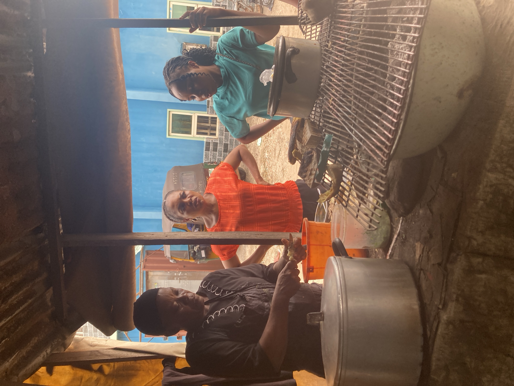 The image captures an outdoor or semi-outdoor food preparation scene, likely a market stall or street vendor in Aba, Nigeria. Three women are visible, engaged in various tasks under a rustic shelter with a corrugated metal roof section visible on the left.
In the foreground, a woman wearing a black long-sleeved top and a dark head covering is seated, attentively peeling or preparing a small root vegetable, possibly ginger, with a knife. A large silver pot with a lid sits beside her. Behind her, another woman in a vibrant orange top stands with hands on her hips, looking upwards towards the camera with a neutral expression.
Further back and to the right, a third woman wearing a teal t-shirt stands, looking down at a large pot or container, possibly stirring its contents or attending to it. She has braids. To her right, a large metal grate or rack is prominent, with various items, including another pot, visible around it, suggesting a cooking or grilling area. An orange bucket is also visible on the ground.
In the background, a bright blue building with white-framed windows stands under what appears to be a clear, sunny daytime sky. Part of a vehicle, possibly a tricycle (keke), is visible between the two standing women, with text "A5500" and "ASOPU" partially legible on its side. The ground appears unpaved, adding to the informal, bustling atmosphere of a street food setup. The overall mood suggests focused activity in a lively environment.