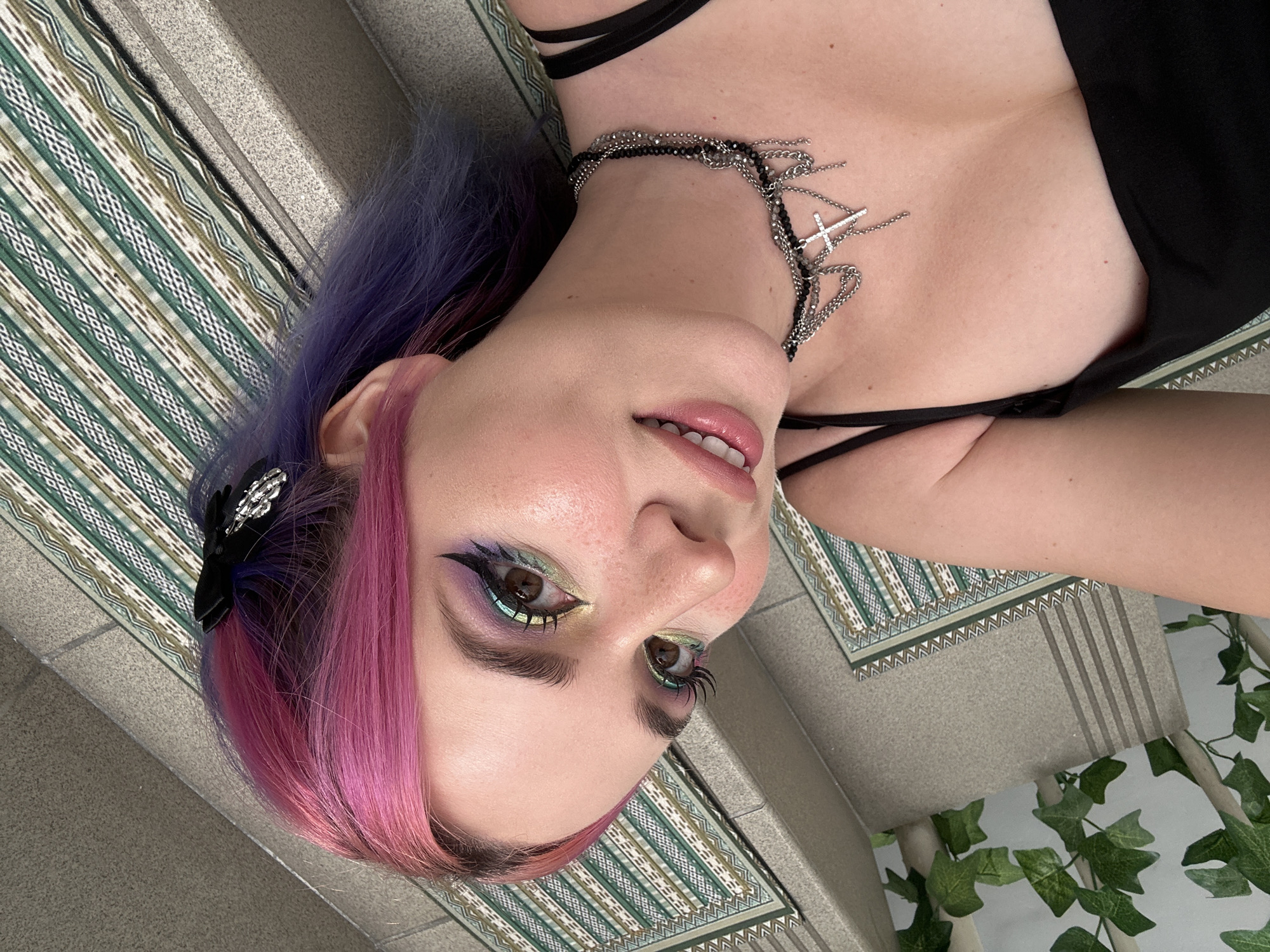 A sideways, eye-level close-up shot features a woman with pink and purple hair and a complex eye makeup look, lying on a textured surface that appears to be a rug or mat. The woman wears a layered black choker necklace with cross charms and a black top. Some artificial green leaves are partially visible on the right side of the frame. The image contains no information about the City of Zagreb, Croatia.