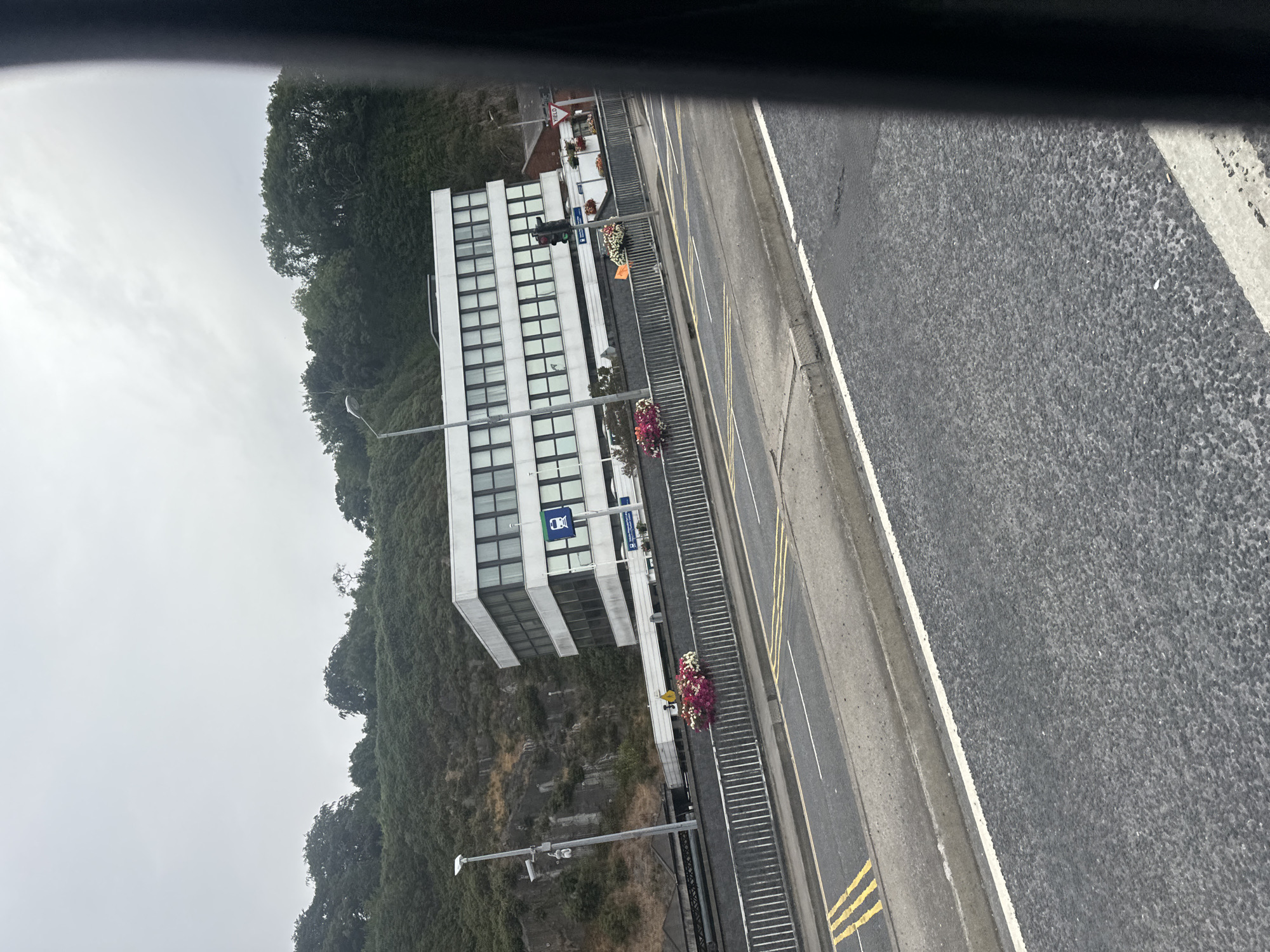 The image shows a modern, multi-story white building with many windows, situated on a hillside with lush green trees. A road with yellow double lines runs in the foreground, with a metal railing separating it from a raised embankment. Flower baskets filled with pink and white flowers are placed along the railing. A street lamp and a CCTV camera are visible on a pole near the building. A sign with a train symbol is also mounted on a pole in front of the building. The scene appears to be a street in Waterford, Ireland, possibly near a train station or public transport hub, given the train signage and the building's architecture. The sky is overcast.