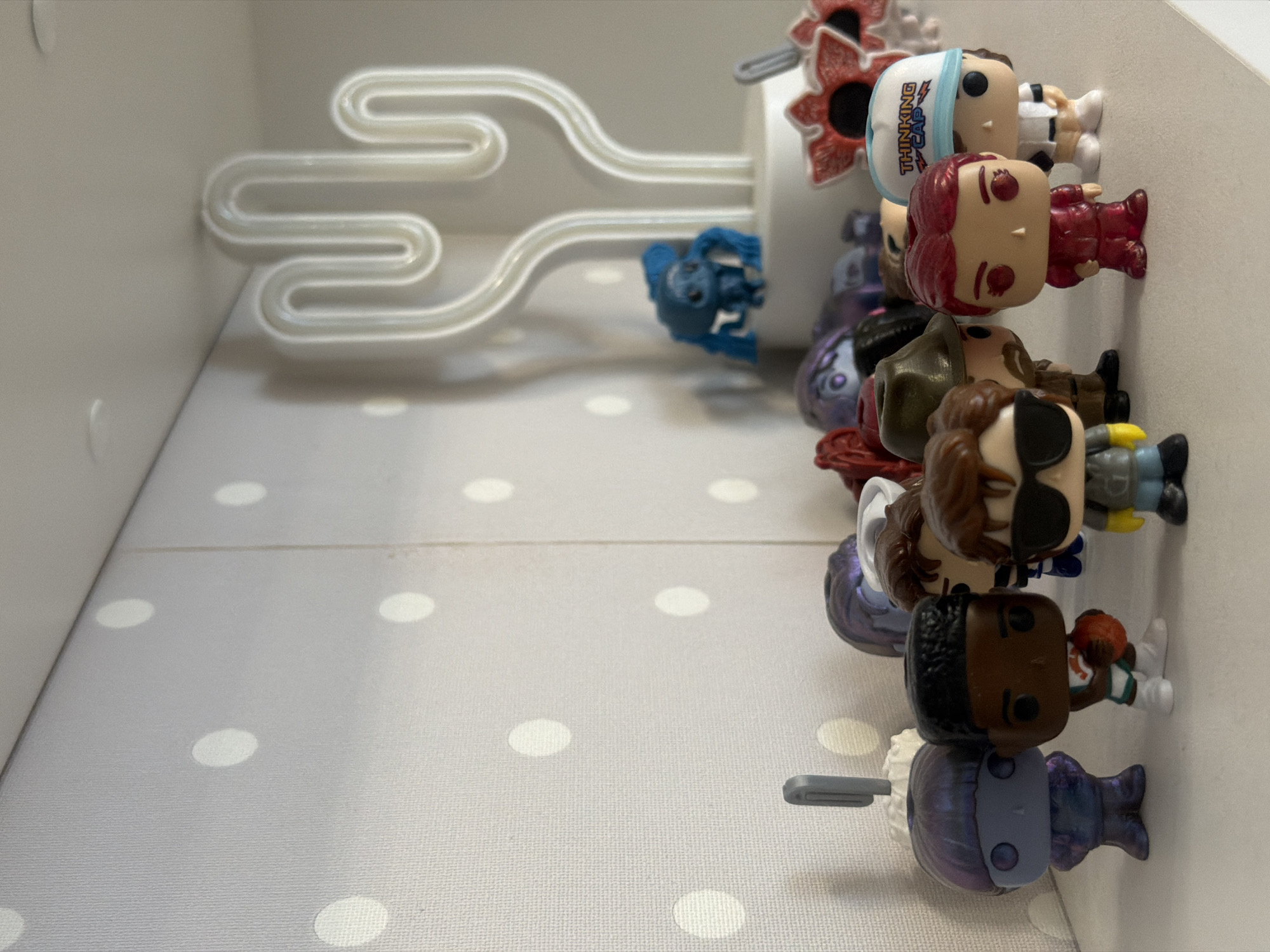 The image shows a shelf containing a white cactus-shaped neon light and a collection of Funko Pop figurines. The shelf has a light gray surface with white polka dots. The Funko Pops are arranged in a row, angled from the right side of the image towards the center. There are approximately eight figurines, each with unique character designs and details. A few of the figurines are partially obscured by others. A small blue figurine stands behind the other figures. The environment appears to be indoors, likely a home or office space. There is no indication of the location being in Rostov-on-Don, Russia.