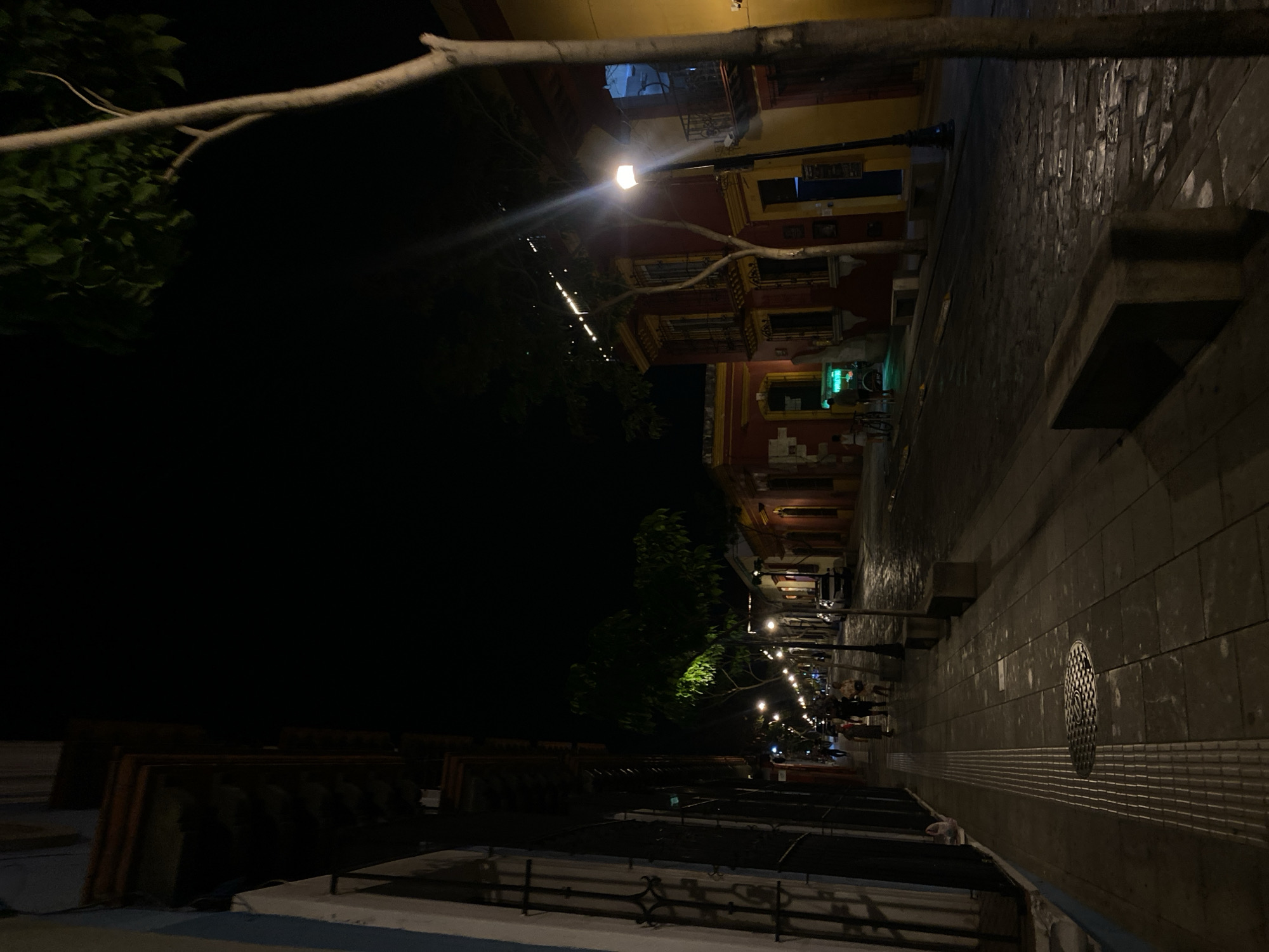 The image depicts a street scene at night in Oaxaca City, Mexico. The buildings are brightly lit with yellow and red paint. Along the sidewalk are lamp posts that illuminate the path. There are several people standing and walking along the street. The foreground of the image shows a series of metal railings.