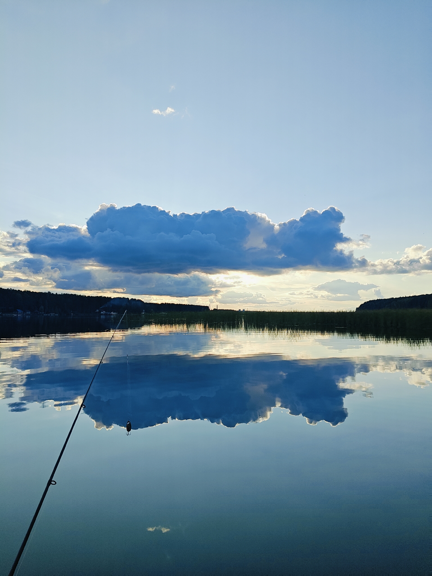 A serene lake scene in Chaykovsky, Russia, under a twilight sky. A fishing rod extends from the lower left corner into the water, its tip just above the surface. A fishing bobber is visible just below the tip. The calm water perfectly reflects the dramatic, cloud-filled sky and the tree-lined horizon. The sun is setting, casting a warm glow through a break in the large, dark clouds. The reflection of the clouds creates a mirrored image on the water's surface, adding to the peaceful atmosphere. The far shore is densely forested, with hints of buildings visible amongst the trees. Tall reeds line the water's edge in some areas. The overall impression is one of tranquility and natural beauty.