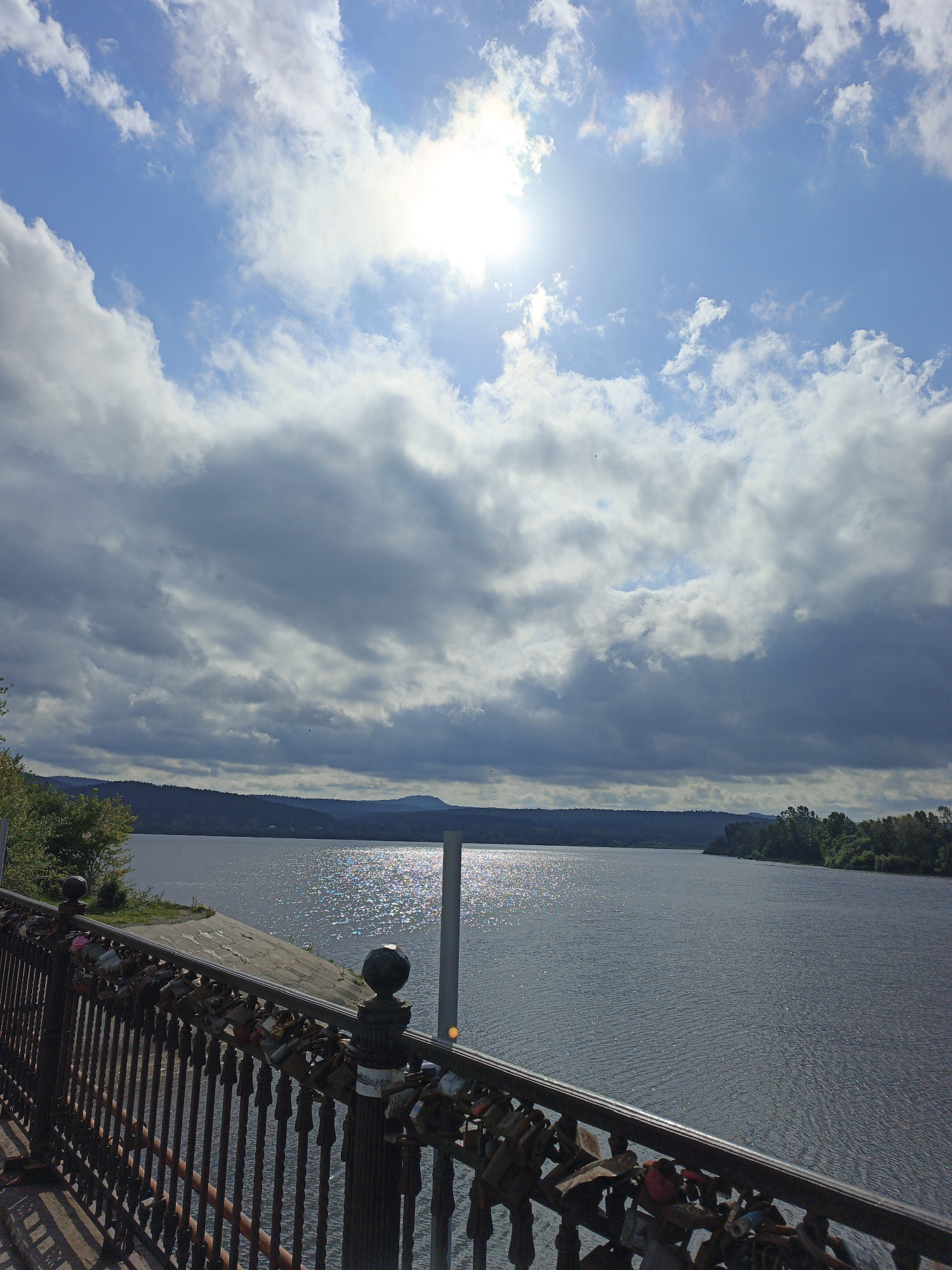 The image captures a scenic view of a body of water, likely a lake or river, under a partly cloudy sky. The sun is visible, casting bright reflections on the water's surface. In the foreground, a dark metal railing of a bridge or promenade is adorned with numerous padlocks, suggesting it is a landmark for couples. Beyond the water, a tree-lined shore and rolling hills form the horizon. The overall impression is one of natural beauty and a touch of romantic symbolism. This scene is located on the waterfront promenade of Zlatoust, Russia.