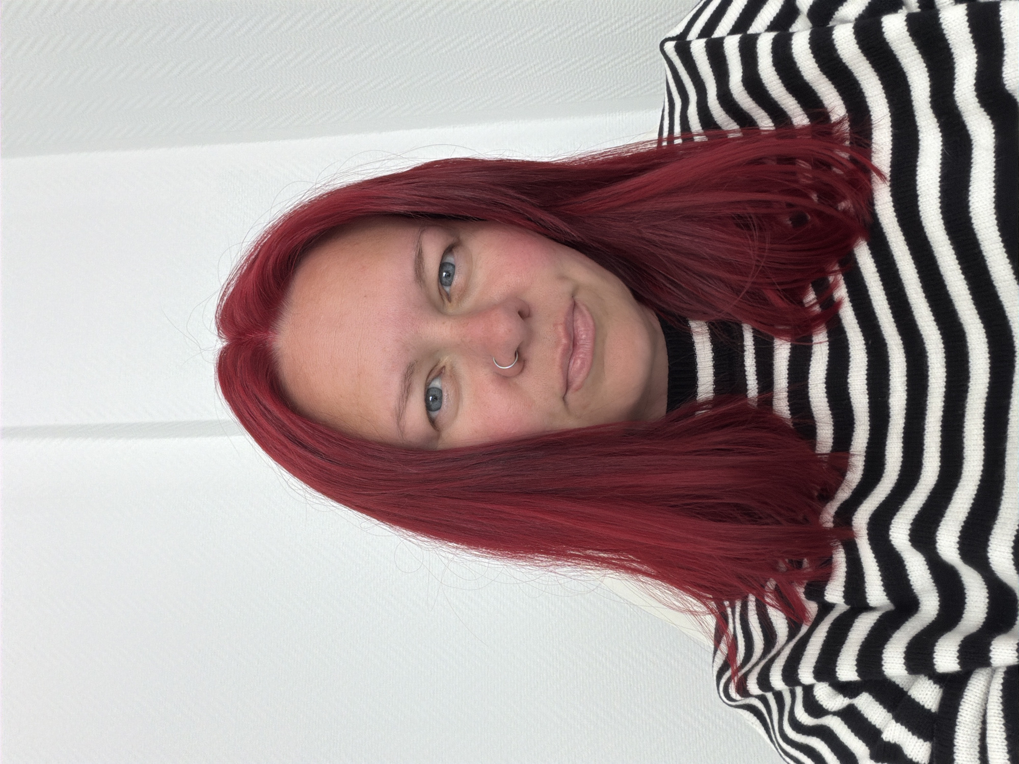 A medium close-up captures a person with fair skin and red hair, wearing a black and white striped sweater. They have blue eyes and a nose ring. The background consists of a white wall with a subtle texture. There are no discernible elements to indicate the location as Chaikovsky, Russia.