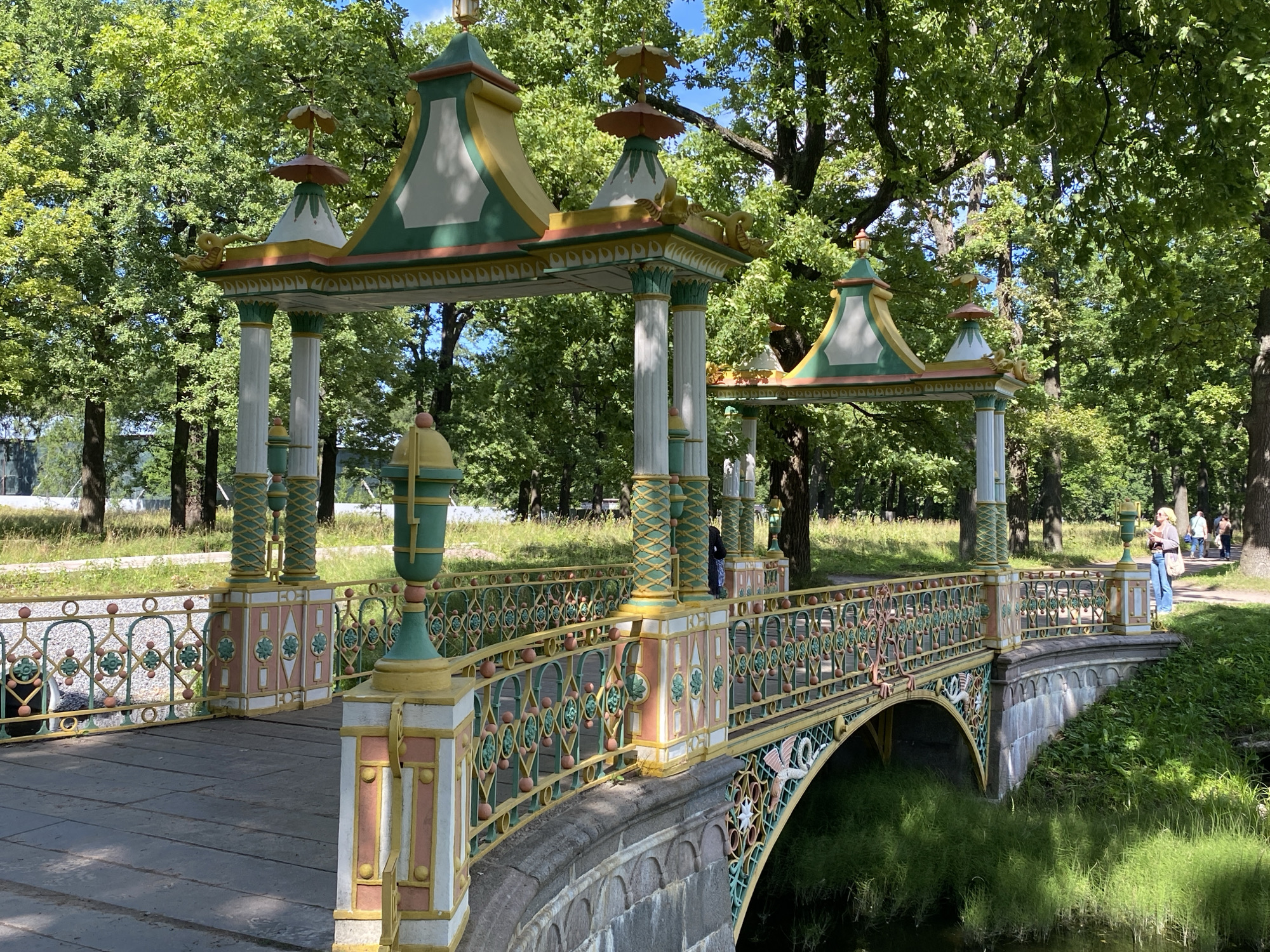 The image depicts a decorative bridge with a pagoda-like roof structure, located in the city of Pushkin, Russia. The bridge, painted in pastel colors of yellow, green, and pink, features ornate railings with intricate patterns and small spherical adornments. Several people are visible on the bridge and in the background, walking along a path and enjoying the park-like setting. Lush green trees and foliage surround the bridge, indicating a natural outdoor environment. The bridge spans a small body of water, likely a canal or pond, with dense green grass growing along its banks. The architecture of the bridge suggests a cultural or historical significance, possibly part of a park or estate.