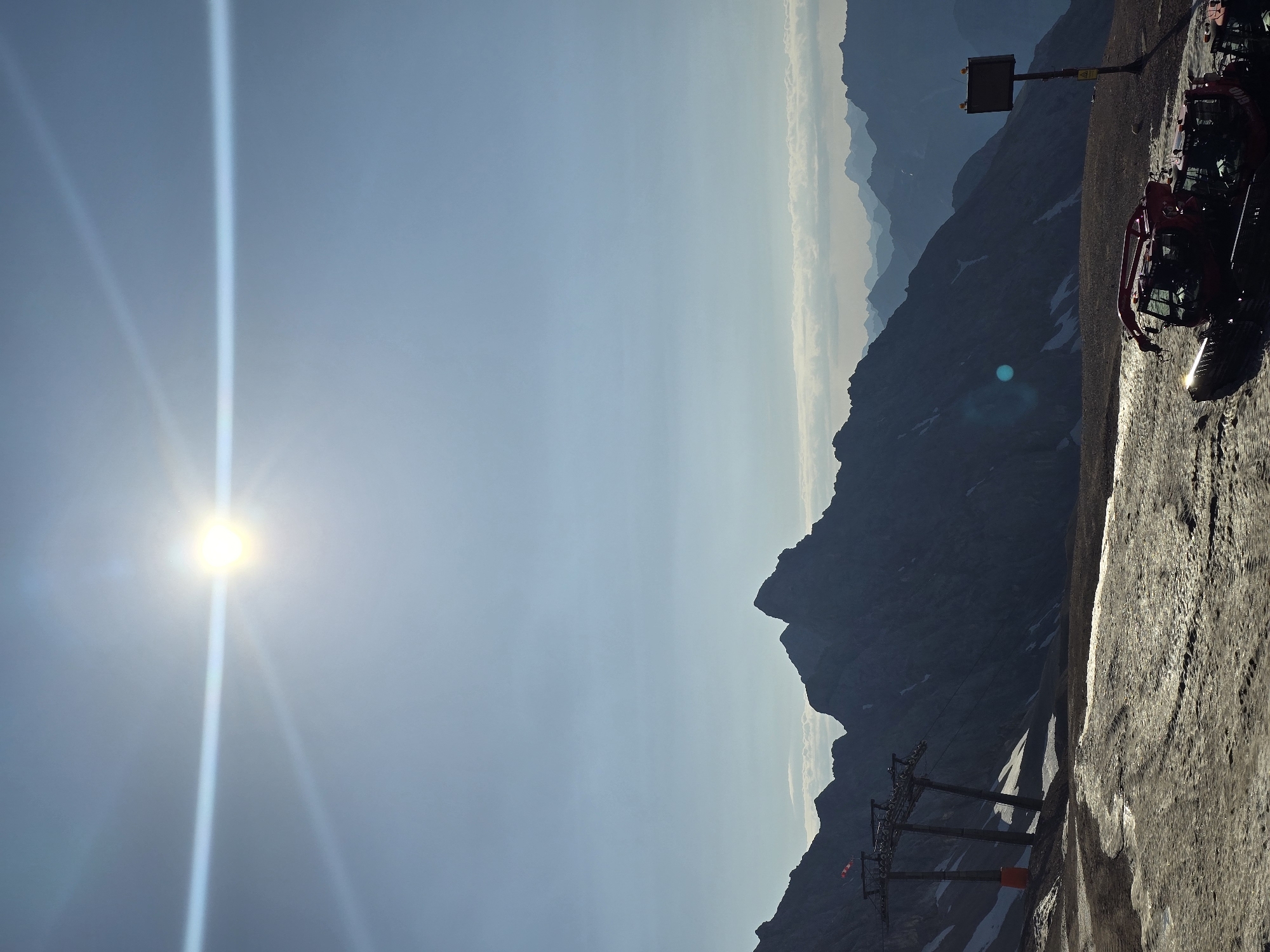 The image depicts a mountain landscape, likely in the Alps near Garmisch-Partenkirchen, Germany, given the presence of ski infrastructure. The sun is high in the sky, creating a lens flare across the upper portion of the image. In the foreground, a red snowcat vehicle is parked on a patch of melting snow and dirt. To the left, a ski lift with support towers is visible, with a red and white flag flying from one of the towers. In the background, a series of majestic, silhouetted mountain ranges stretch across the horizon under a clear blue sky. The overall scene suggests a high-altitude location, possibly a ski resort during the off-season or early morning/late evening.