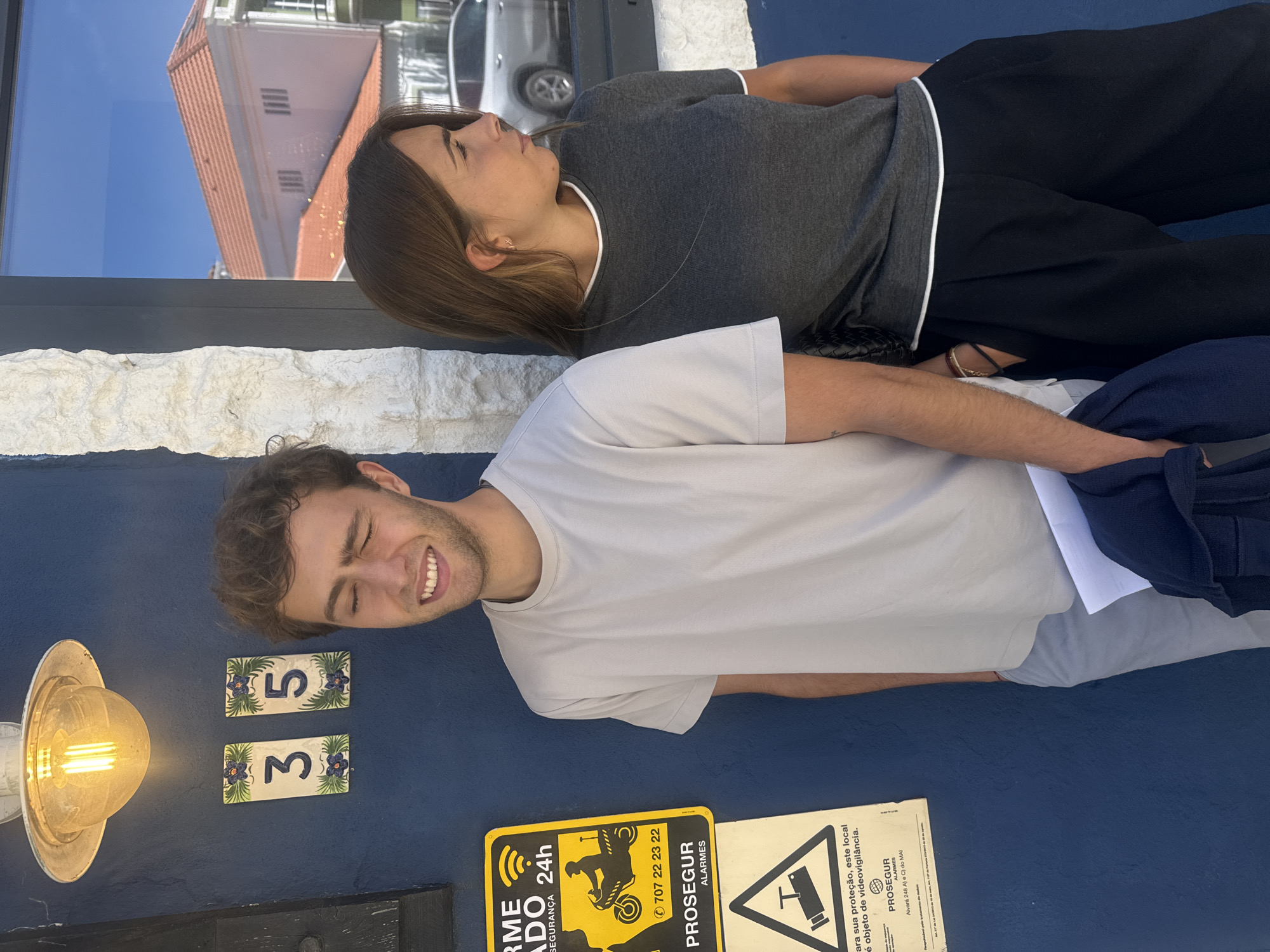 An outdoor portrait captures two individuals standing against a vibrant blue building facade under bright daylight, likely in Torres Vedras, Portugal, given the visible Portuguese text. The man in the foreground, wearing a light-colored t-shirt, smiles broadly with his eyes closed, showing a joyful expression. He has short, curly brown hair and light stubble. His left arm is visible, draped with a dark blue or purple item.
Behind him, a woman with long brown hair wears a grey t-shirt with white trim and black pants. Her eyes are also closed, and her expression appears serene and peaceful.
The blue facade features a metallic outdoor light fixture with an Edison-style bulb, and two decorative white tiles with blue floral designs, displaying the numbers "3" and "5". Below these, two security signs are mounted. The yellow sign shows a motorcycle icon and reads "ALARME ADO 24h SEGURANÇA", with a phone number "707 22 23 22" and "PROSEGUR ALARMES". The white sign states, "Para sua proteção, este local é objeto de videovigilância.", also featuring "PROSEGUR ALARMES" and "Alvará 248 A) C) do MAI", alongside a CCTV camera icon.
Above the blue wall, a textured white stone or stucco wall separates it from a large window reflecting a bright blue sky and the upper part of a building with a distinct red tiled roof. A partial reflection of a silver car is also visible in the window. The overall scene suggests a sunny day, with the individuals enjoying the light or posing for a photo.
