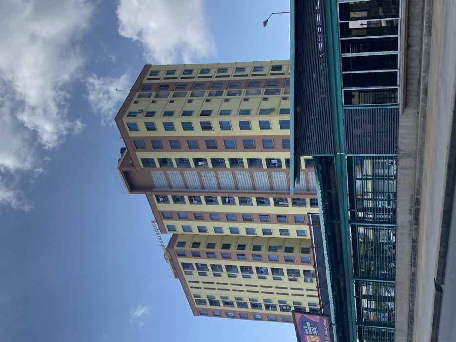 The image displays a tall, multi-story building dominating the left and central parts of the frame, set against a bright blue sky dotted with scattered white clouds. The building has a distinctive architectural style, featuring light yellow walls accented by horizontal brown bands that separate its many floors. Numerous rectangular windows, appearing dark or blue, are visible across the facade, with some balconies showing clothes hanging to dry, suggesting residential use. Partial text on the side of the building appears to read "Bird Residences."
In the foreground, on the right side, an elevated infrastructure component, possibly an overpass, walkway, or part of a public transport station, extends into the frame. This structure is built from concrete and features prominent green metal railings and dark slatted panels. A street light pole is visible above the building. At the bottom right, a small portion of a billboard is visible, displaying text that includes "20" and "EA CLC official."
The scene depicts a clear, sunny daytime. No specific human activity is actively taking place. The overall impression is that of an urban landscape, consistent with the provided context of Dar es Salaam, Tanzania, featuring modern architecture and infrastructure.