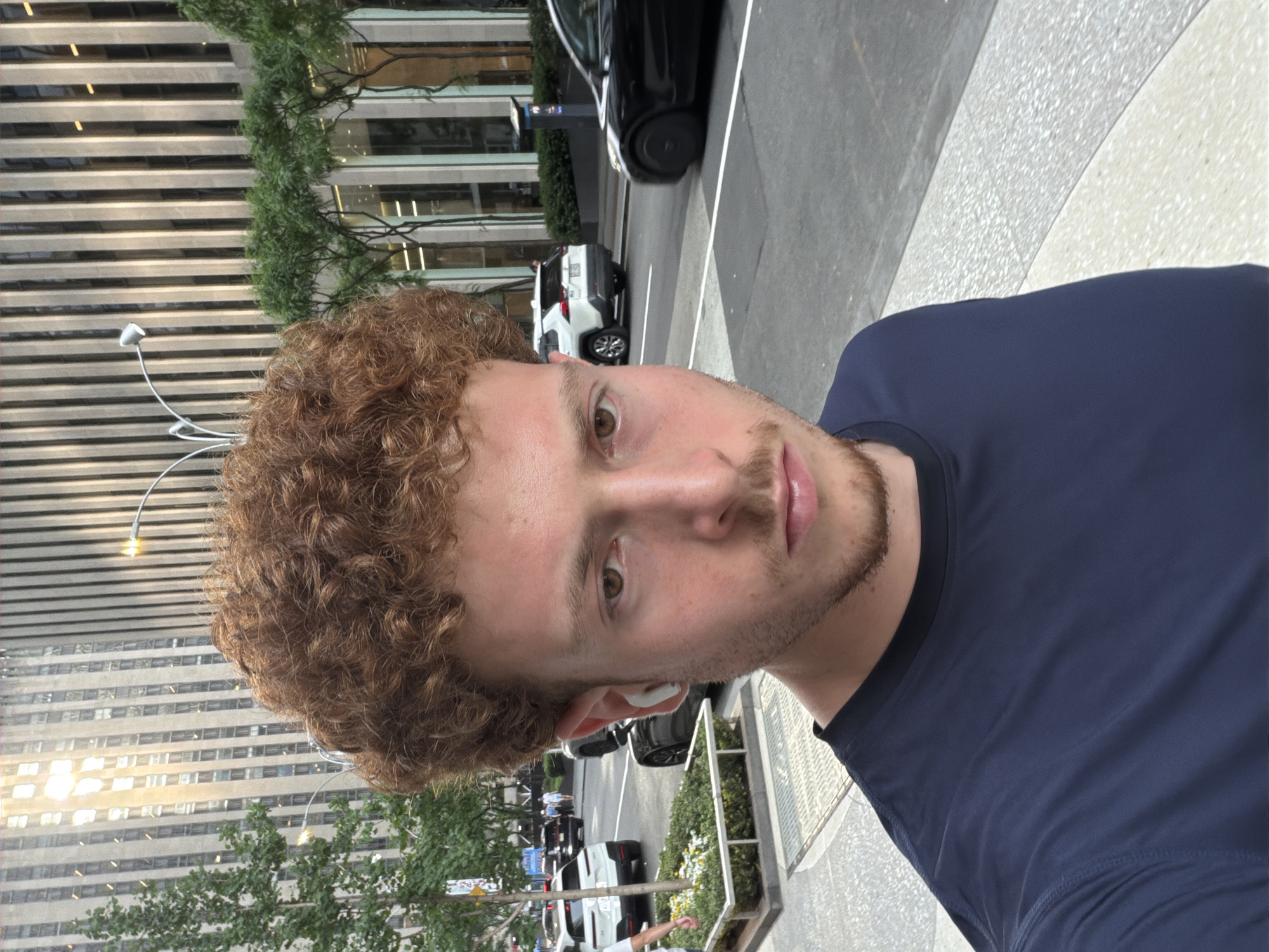 A light-skinned male with curly reddish-brown hair, a short beard, and wearing a dark blue shirt, stands in an urban environment. He has an earbud in his left ear. Behind him is a city street with cars parked and moving. Further in the background, a tall building with a pattern of horizontal stripes is visible, along with trees. The location appears to be in New York City, based on the architecture and urban setting.