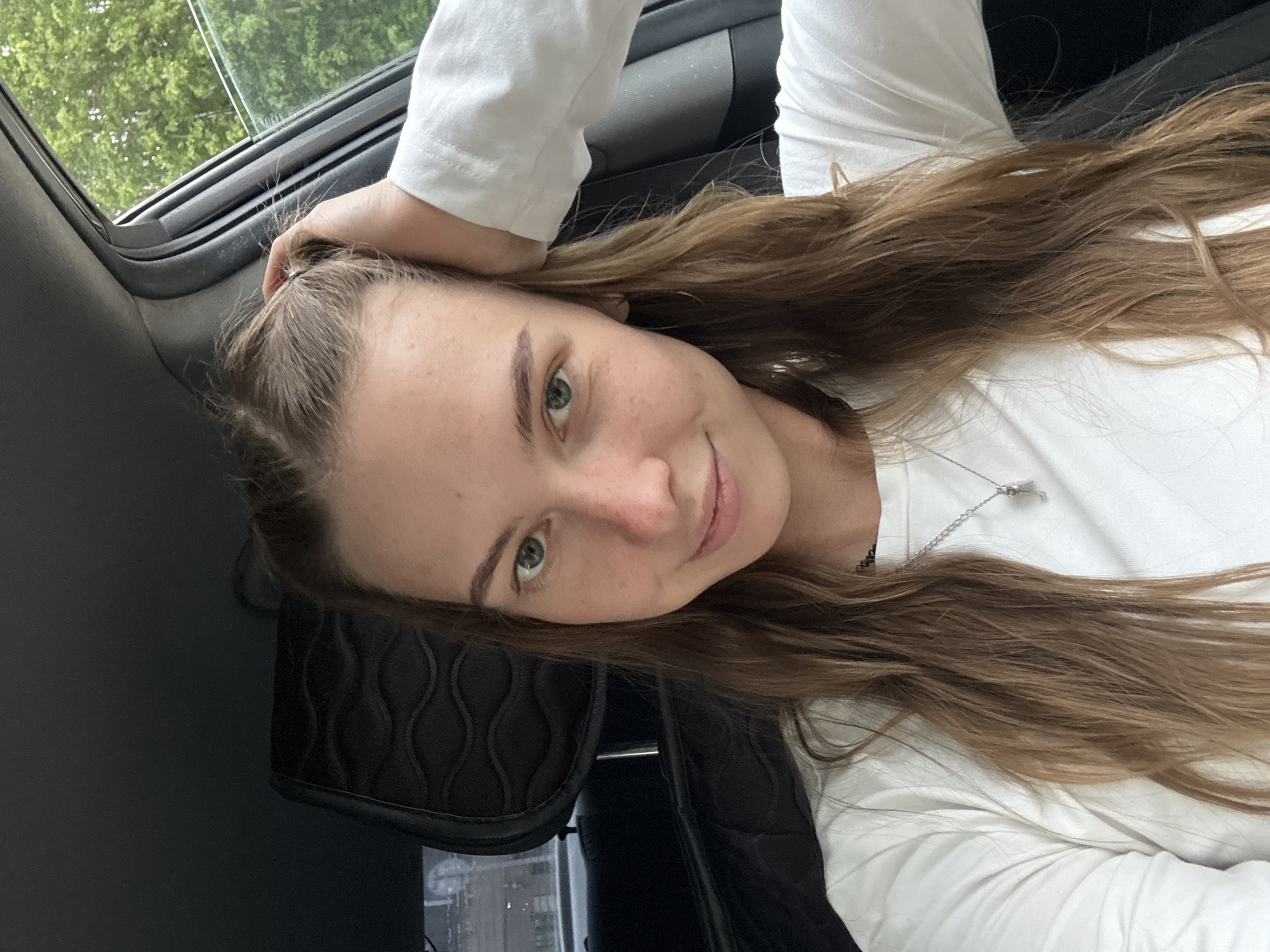 A young woman with long, light brown hair pulled back with a hand is seated in the driver's seat of a car. She is wearing a white long-sleeved shirt and a silver necklace with a key pendant. Her gaze is directed at the camera, and she has a slight smile on her face. The interior of the car is visible, including a black headrest with a patterned design. Outside the car window, green trees and foliage can be seen, indicating an outdoor setting, possibly on the outskirts of Perm, Russia, during daylight hours.