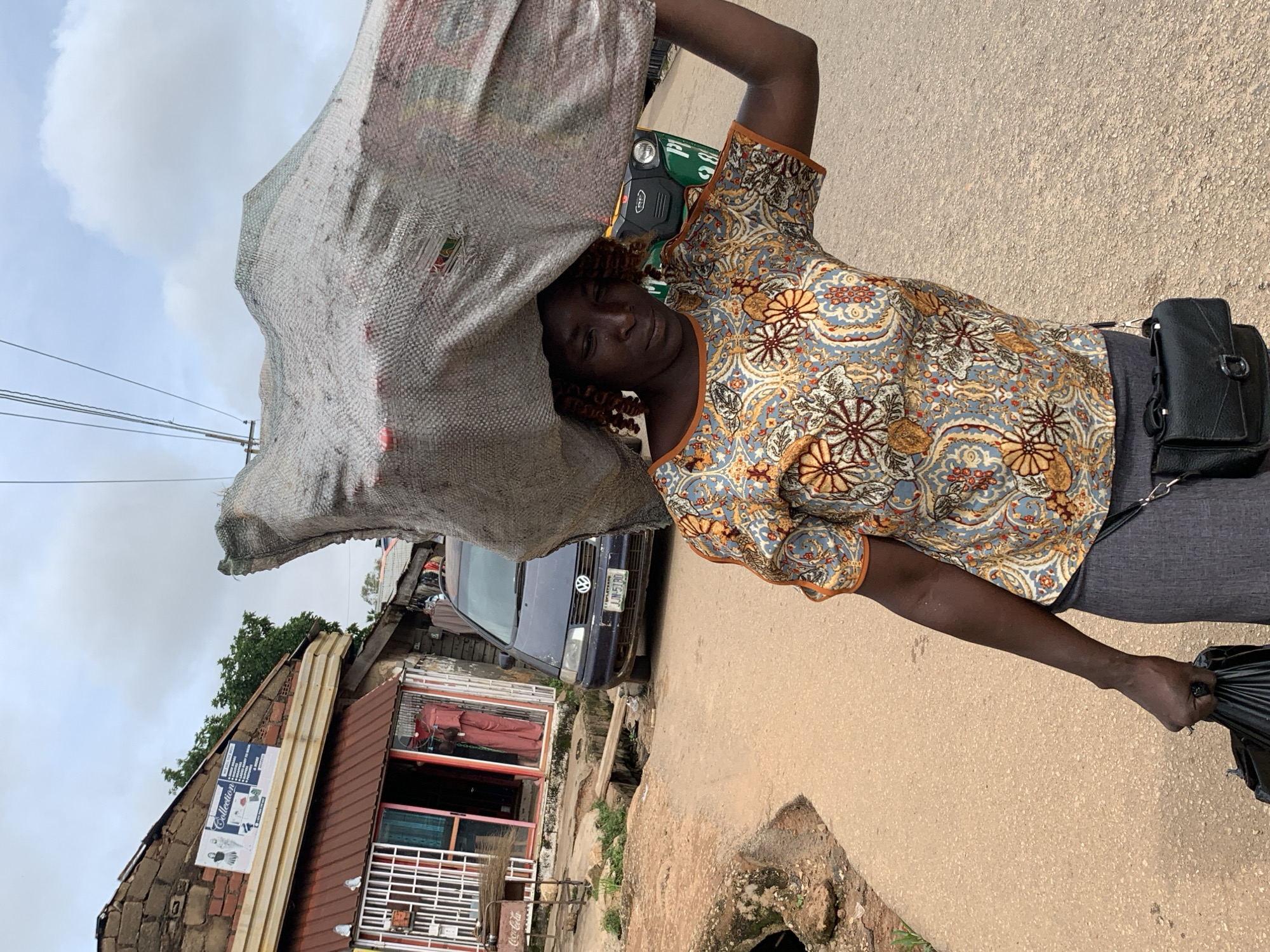 A woman carries a large, gray, netted bag filled with goods on her head, walking on a paved road in Gura Top, Nigeria. She wears a colorful, patterned short-sleeved shirt and a gray skirt, with a black purse slung across her body. Her right hand holds a black plastic bag. In the background, a dark blue Volkswagen car is parked on the side of the road, and a green and yellow auto-rickshaw is visible further down. Shops with awnings and displays line the left side of the road. The sky is overcast with scattered clouds.