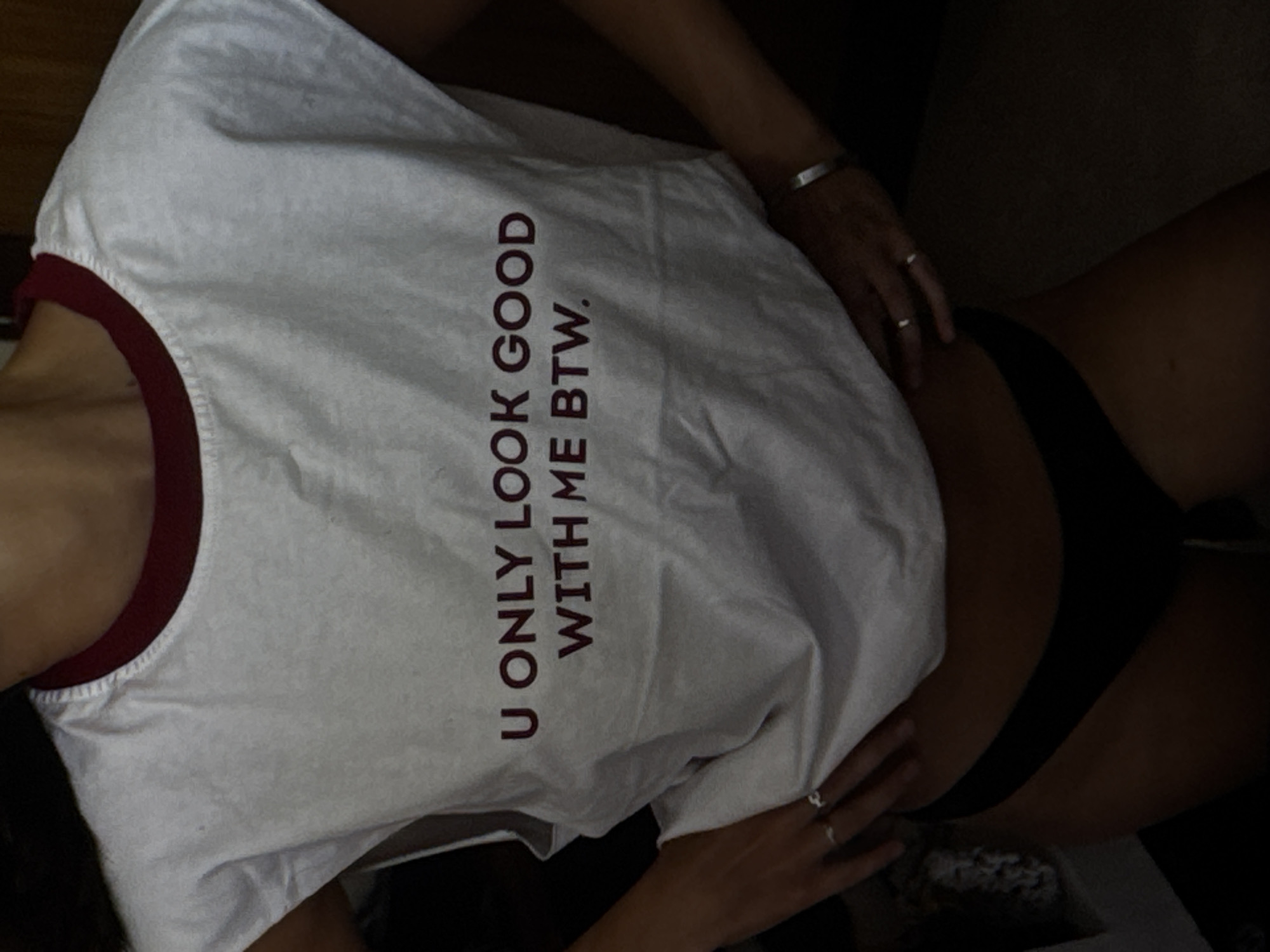 A person is posing for a photo wearing a white t-shirt with burgundy lettering that reads "U ONLY LOOK GOOD WITH ME BTW." The t-shirt has a burgundy trim around the neckline. The person is wearing black underwear or bikini bottoms. The person is wearing two rings on their left hand and a bracelet on their right wrist. The background is dark and out of focus, but appears to be an indoor setting. The lighting is dim. The image does not contain any specific features that would allow for the determination of its location in Kuta Selatan, Indonesia.