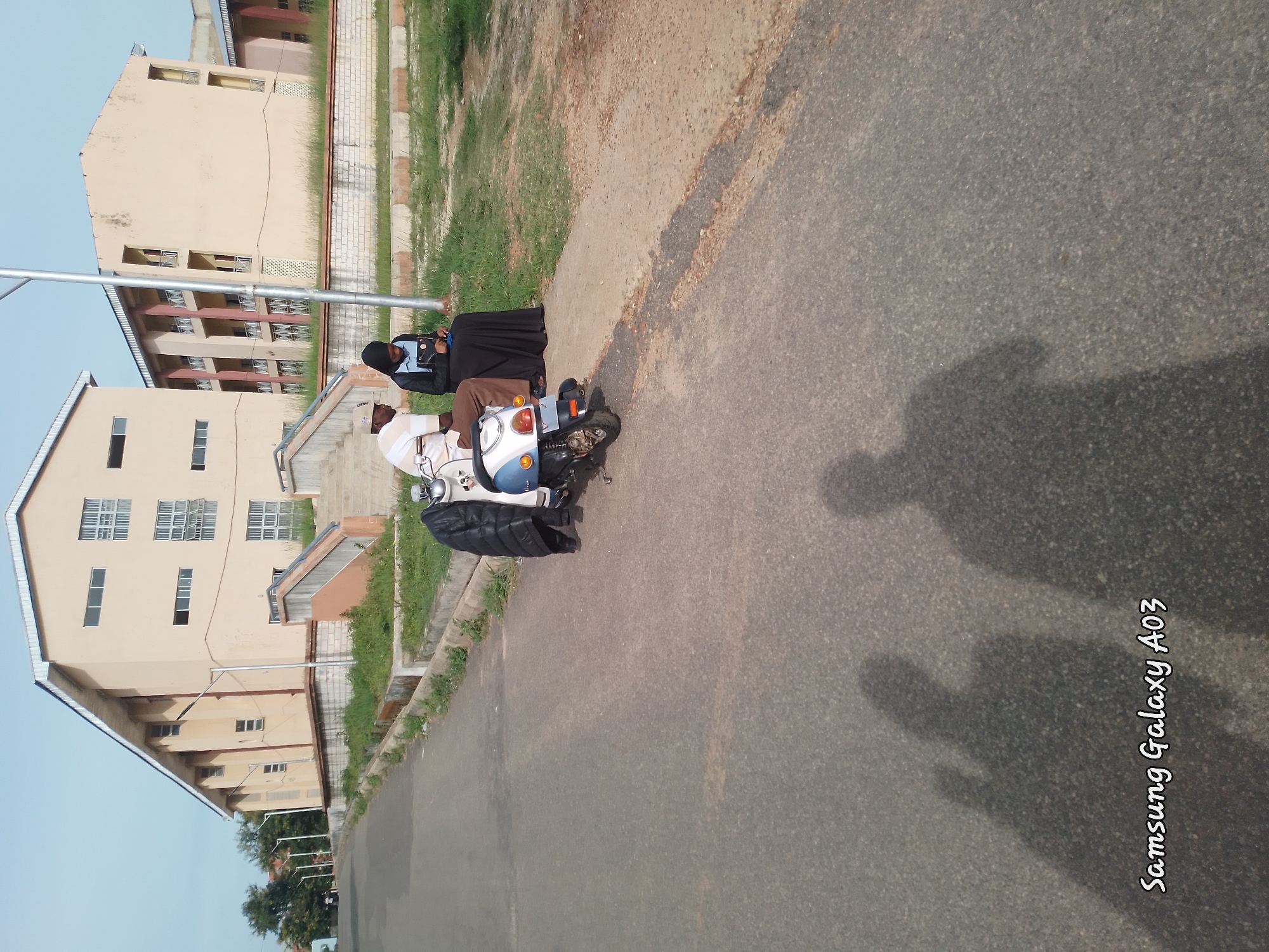 Two individuals are seated next to a parked scooter on an asphalt road in front of buildings. One person, a male wearing a light-colored striped shirt and a cap, is seated on the scooter. The other person, a female wearing a black hijab and a dark dress, is seated on the ground next to him. The scooter is light blue and white. In the background, there are two multi-story buildings with light-colored exteriors and windows. The environment appears to be an outdoor setting, possibly a campus or public area, with some greenery and paved walkways. The scene is captured under natural lighting, casting shadows on the road. The text "Samsung Galaxy A03" is present in the lower left corner of the image.