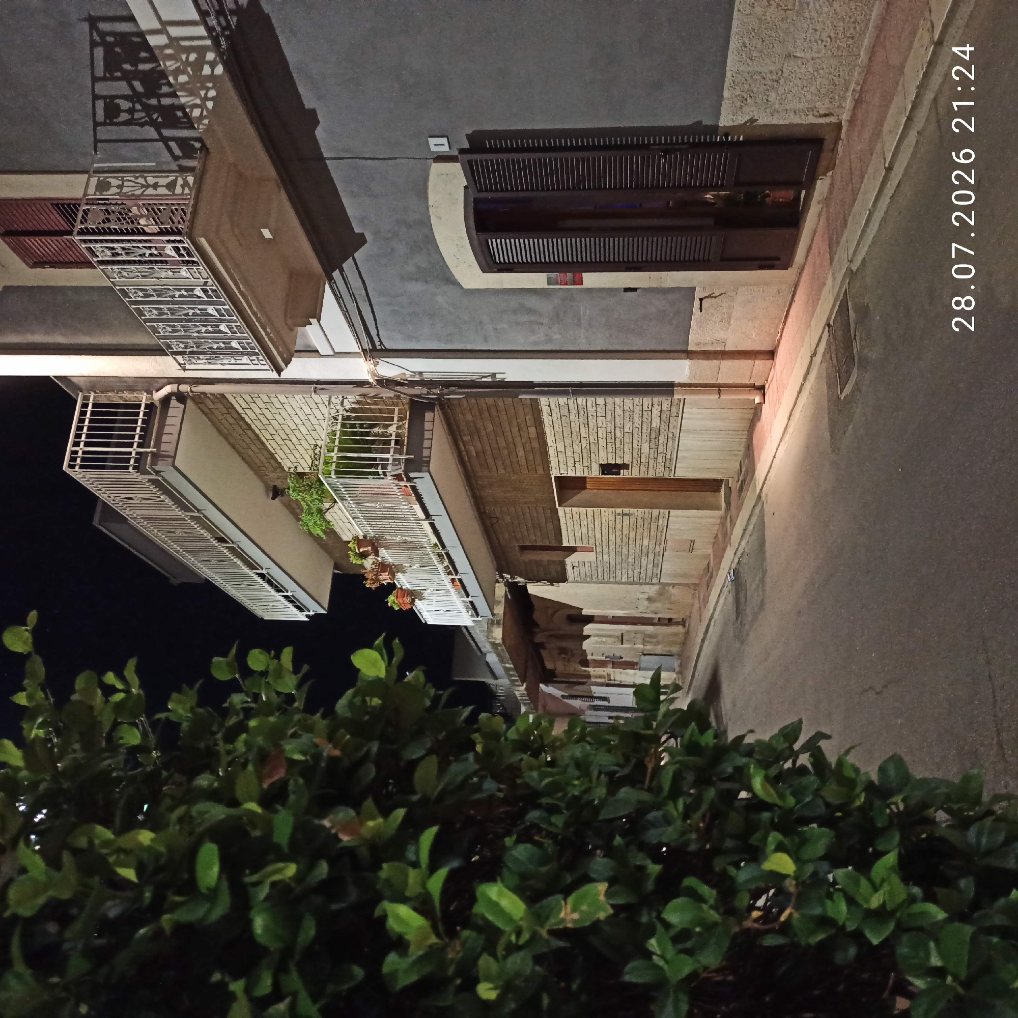 The image depicts an outdoor scene at night, likely in an urban or semi-urban setting in Bitonto, Italy. The perspective is looking upwards, at an angle, towards the facades of buildings and a street. In the foreground, a lush, dark green bush or hedge takes up the lower portion of the frame, obscuring the very bottom.
Dominating the mid-ground and background are the sides of buildings with multiple stories. These buildings feature balconies with decorative metal railings. One balcony has potted plants with red-brown blooms. Another balcony above it appears to have a dark red metal shutter. The building walls are a mix of smooth gray surfaces and textured, light-colored brickwork. Above the balconies, windows are visible, some with dark, slatted shutters, suggesting a residential or commercial structure. A narrow paved street or sidewalk runs along the base of the buildings, illuminated by a warm light source from above. Shadows are cast by the balconies and architectural elements onto the building facades.
The time of day is clearly night, as indicated by the darkness of the sky and the artificial lighting. The weather appears calm. A timestamp is visible in white text, vertically oriented on the right side of the image, reading "28.07.2026 21:24". This suggests the photograph was taken on July 28, 2026, in the evening. There are no people visible in the scene, and the overall atmosphere is quiet and still. The focus is on the architecture and the surrounding foliage.