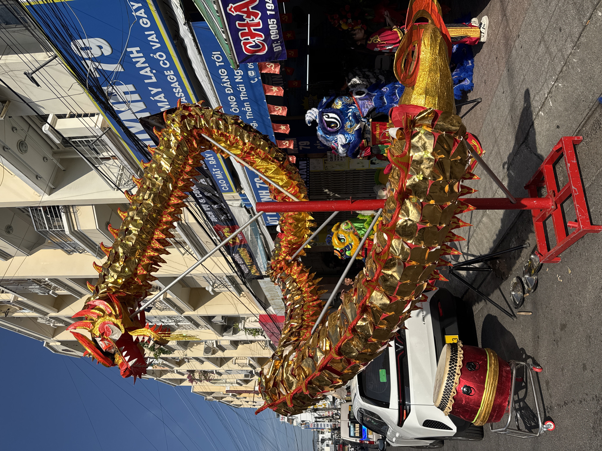 This image depicts a street scene in Nha Trang Ward, Vietnam, likely set up for a celebration or performance. In the foreground, a large, golden dragon prop, adorned with red accents, is prominently displayed on a red stand. The dragon's head is detailed with red and gold scales, sharp teeth, and expressive eyes. To the left of the dragon, a red drum on a metal cart with wheels is visible, along with some cymbals and drumsticks on the ground.
Behind the dragon, a white car is parked, and beyond that, a row of buildings lines the street. These buildings appear to be multi-story with balconies and air conditioning units. Several shops are visible at street level, indicated by signs with Vietnamese text. One prominent blue sign reads "MÁY LẠNH 79" and "MASSAGE CỔ VAI GÁY," suggesting an air conditioning shop and a massage service. Other signs display information about services and promotions.
Several individuals are present in the scene. Two people are dressed in vibrant blue and green lion dance costumes, with detailed masks and furry bodies. One person in a red and gold outfit, possibly a performer, stands near the right side of the frame. Other individuals are partially visible in the background, near the shops. The environment is an urban street with clear blue skies overhead, casting shadows from the dragon and other objects onto the pavement. The overall impression is one of a lively, culturally significant event taking place in a Vietnamese city.